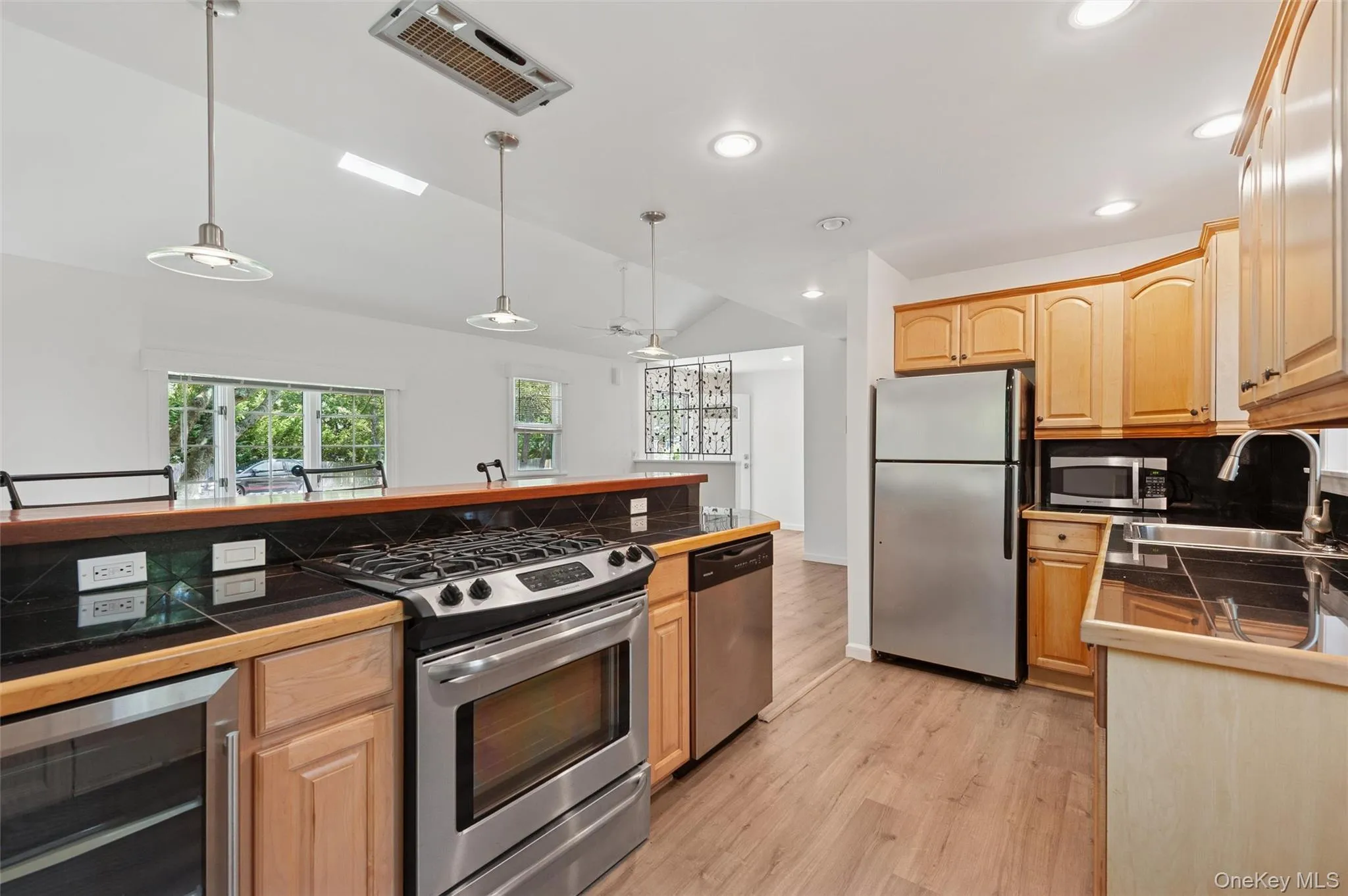 Kitchen featuring appliances with stainless steel finishes, lofted ceiling, beverage cooler, light wood finished floors, and light brown cabinetry Kitchen featuring appliances with stainless steel finishes, lofted ceiling, beverage cooler, light wood finished floors, and light brown cabinetry