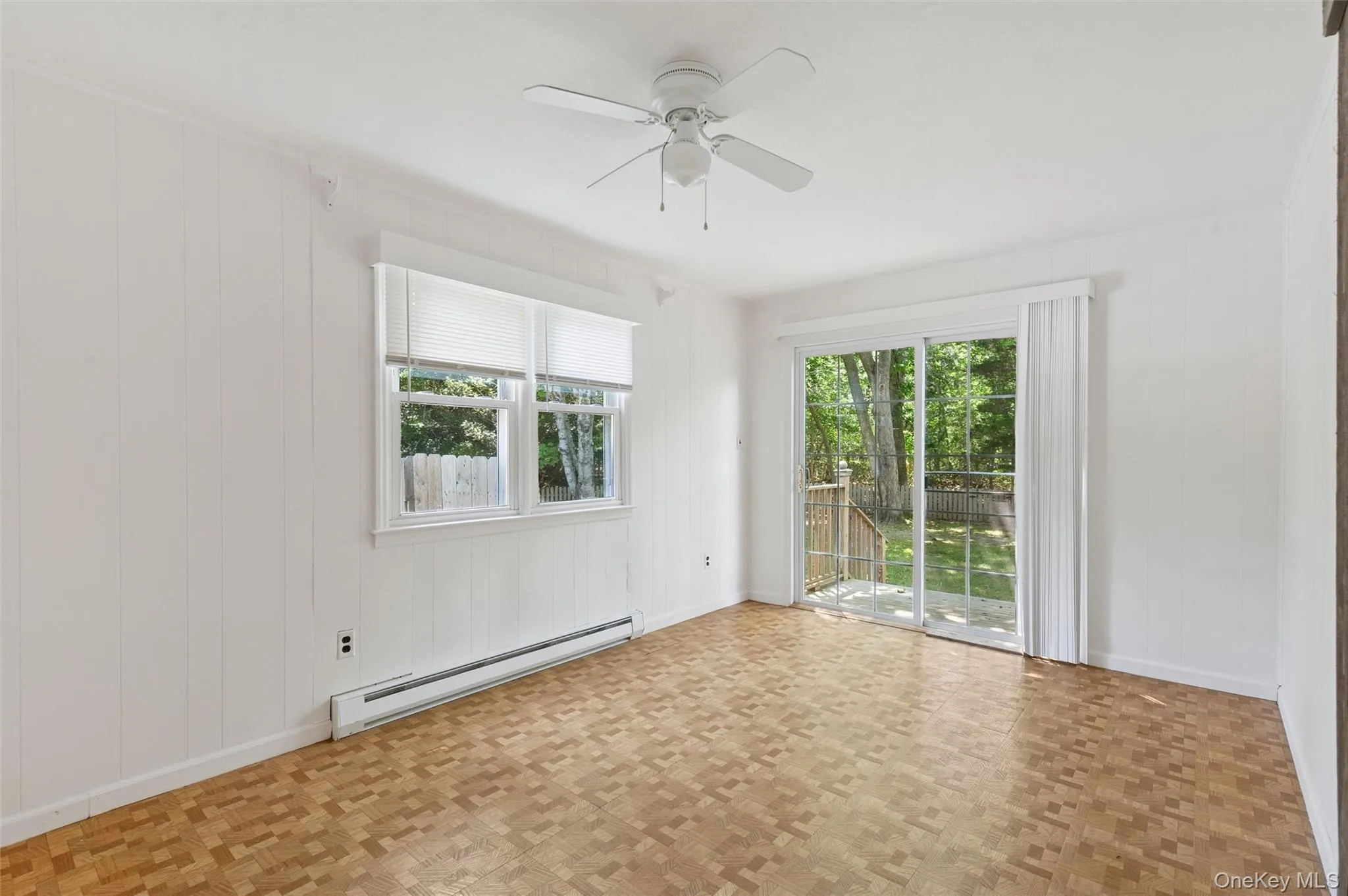 Empty room featuring baseboard heating, plenty of natural light, a ceiling fan, wood walls, and parquet flooring Empty room featuring baseboard heating, plenty of natural light, a ceiling fan, wood walls, and parquet flooring