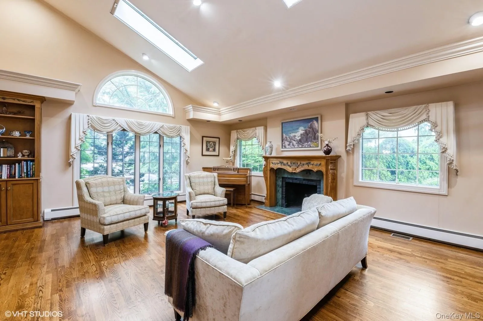 Living room with high vaulted ceiling, a skylight, light wood finished floors, a premium fireplace, and a baseboard radiator Living room with high vaulted ceiling, a skylight, light wood finished floors, a premium fireplace, and a baseboard radiator