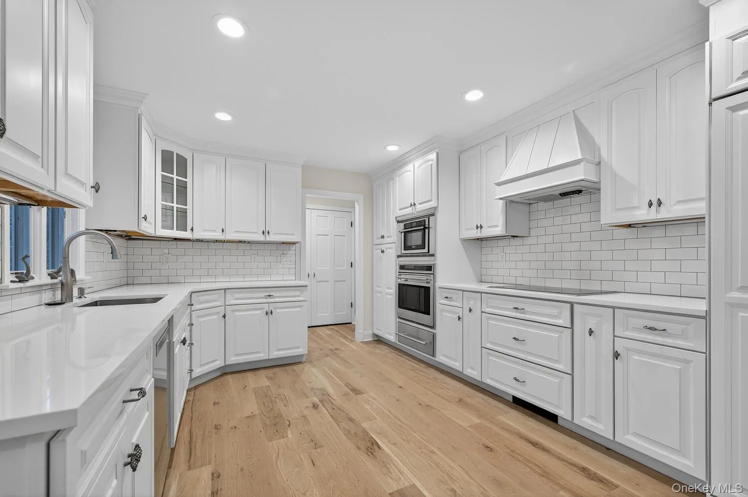 Kitchen featuring white cabinetry, glass insert cabinets, custom range hood, light wood finished floors, and stainless steel appliances Kitchen featuring white cabinetry, glass insert cabinets, custom range hood, light wood finished floors, and stainless steel appliances