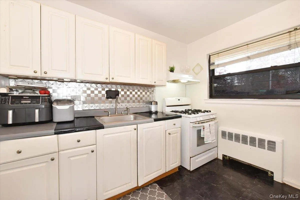 Kitchen featuring white gas range oven, radiator, dark countertops, backsplash, and under cabinet range hood Kitchen featuring white gas range oven, radiator, dark countertops, backsplash, and under cabinet range hood