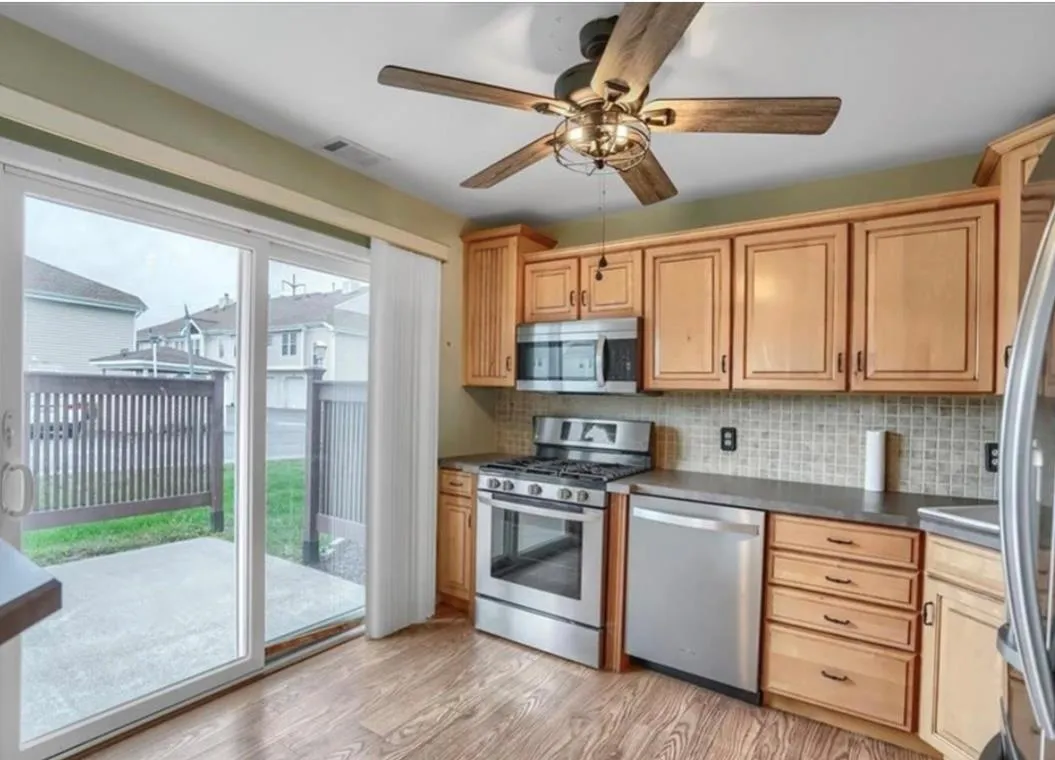 Kitchen featuring appliances with stainless steel finishes, light wood-style flooring, dark countertops, tasteful backsplash, and a ceiling fan Kitchen featuring appliances with stainless steel finishes, light wood-style flooring, dark countertops, tasteful backsplash, and a ceiling fan
