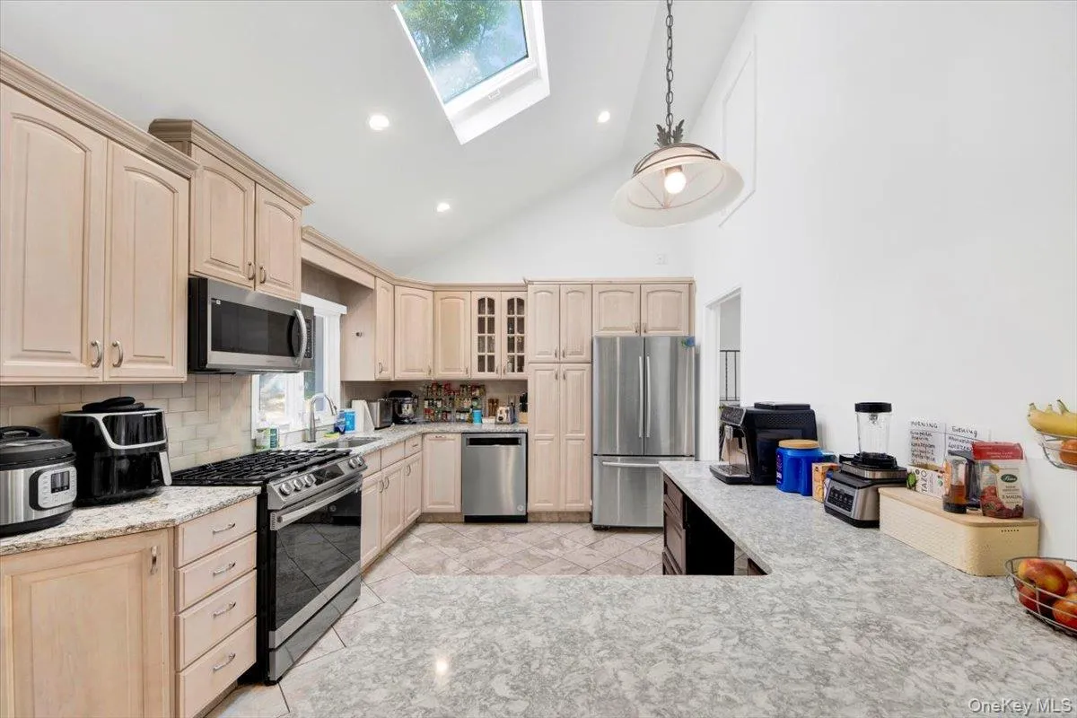Kitchen with light brown cabinetry, a skylight, appliances with stainless steel finishes, high vaulted ceiling, and glass insert cabinets Kitchen with light brown cabinetry, a skylight, appliances with stainless steel finishes, high vaulted ceiling, and glass insert cabinets