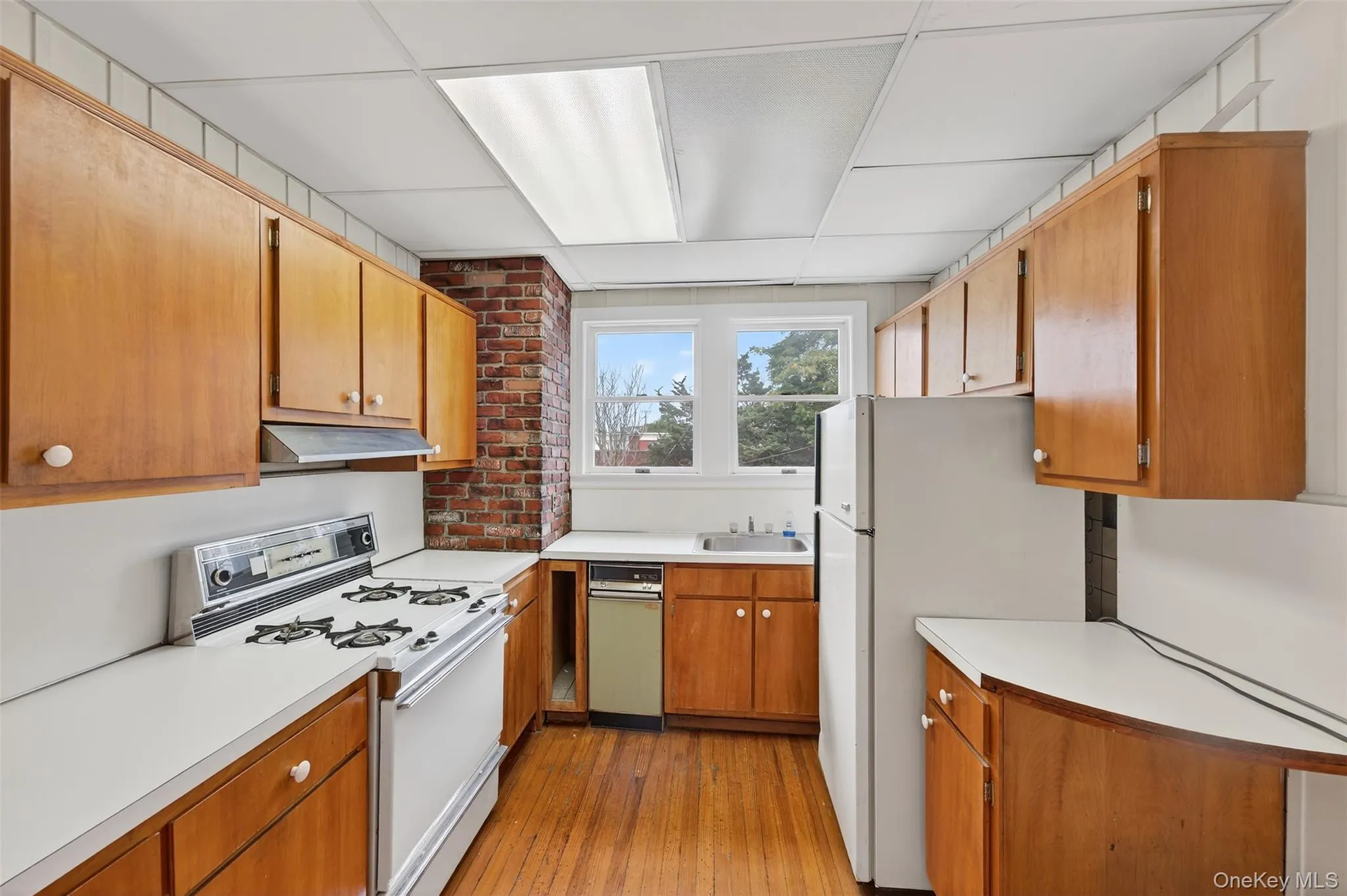Kitchen featuring white appliances, light countertops, light wood-style flooring, a drop ceiling, and brown cabinets Kitchen featuring white appliances, light countertops, light wood-style flooring, a drop ceiling, and brown cabinets