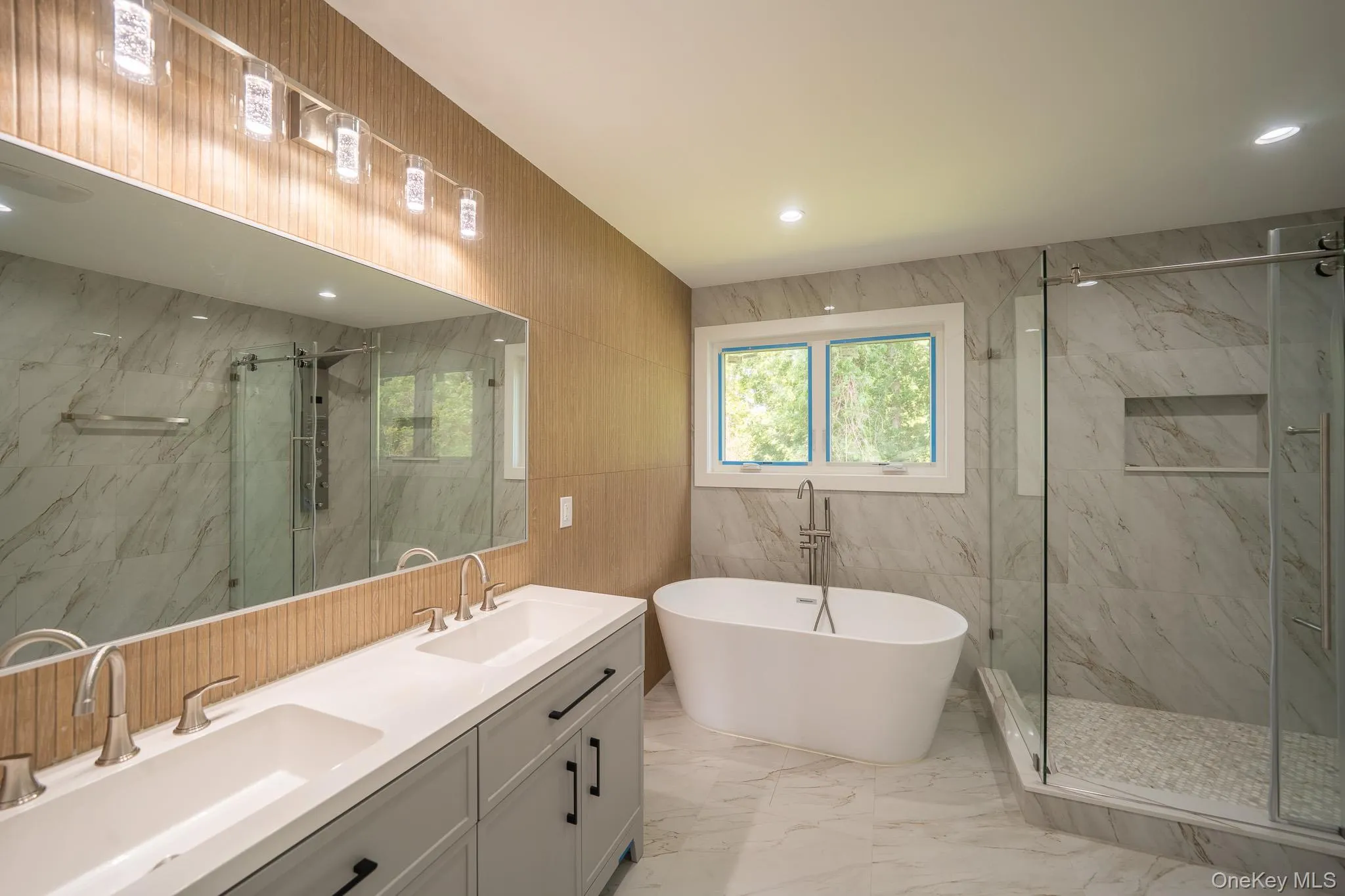 Full bath featuring tile walls, marble look tile flooring, a marble finish shower, a freestanding tub, and double vanity Full bath featuring tile walls, marble look tile flooring, a marble finish shower, a freestanding tub, and double vanity