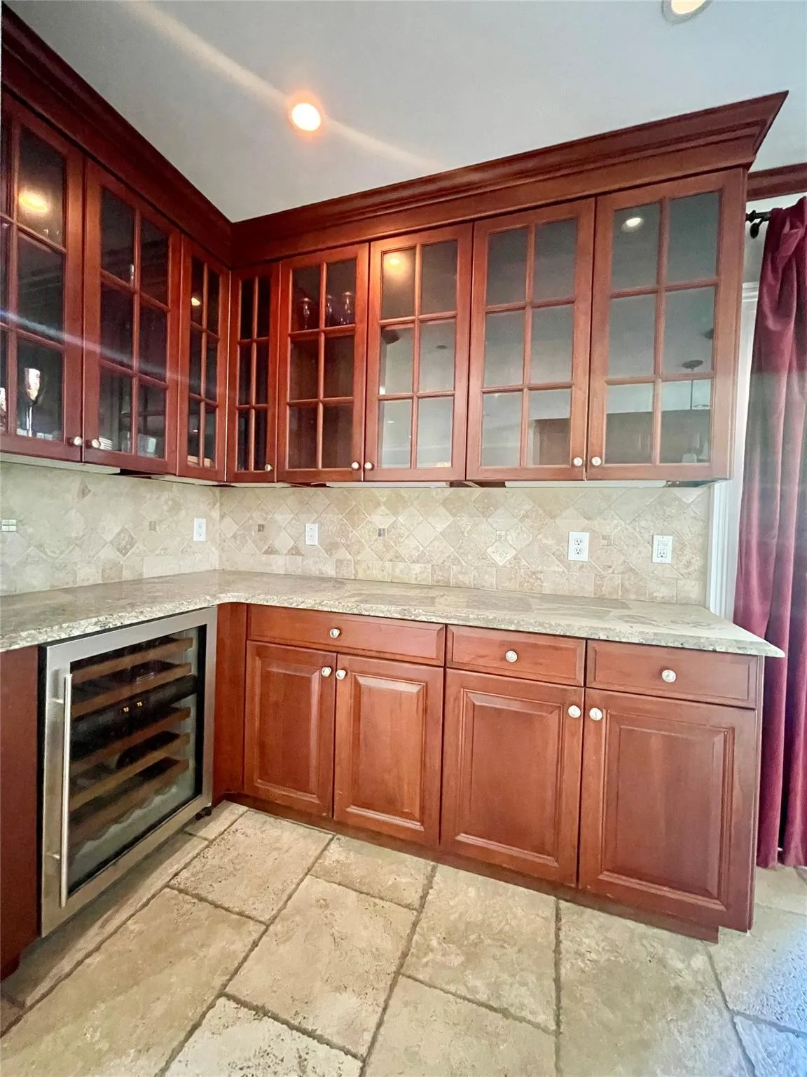 Kitchen with beverage cooler, reddish brown cabinets, stone tile floors, light stone counters, and recessed lighting Kitchen with beverage cooler, reddish brown cabinets, stone tile floors, light stone counters, and recessed lighting