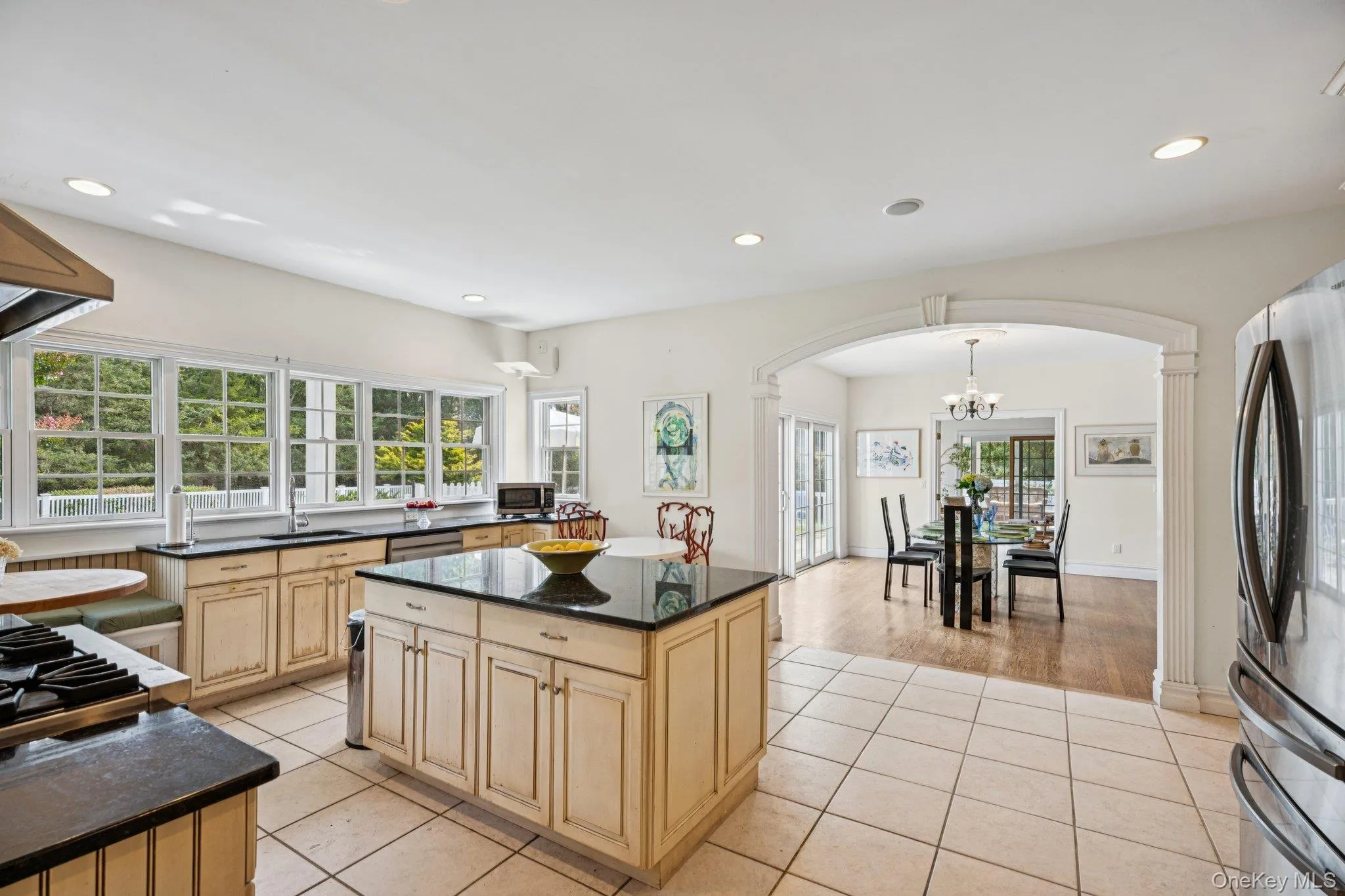 Gourmet kitchen showcasing granite countertops, a generous island, custom cabinetry with glass inserts, an inviting eat-in area, recessed lighting, and light-patterned tile flooring illuminated by an abundance of natural light. Gourmet kitchen showcasing granite countertops, a generous island, custom cabinetry with glass inserts, an inviting eat-in area, recessed lighting, and light-patterned tile flooring illuminated by an abundance of natural light.