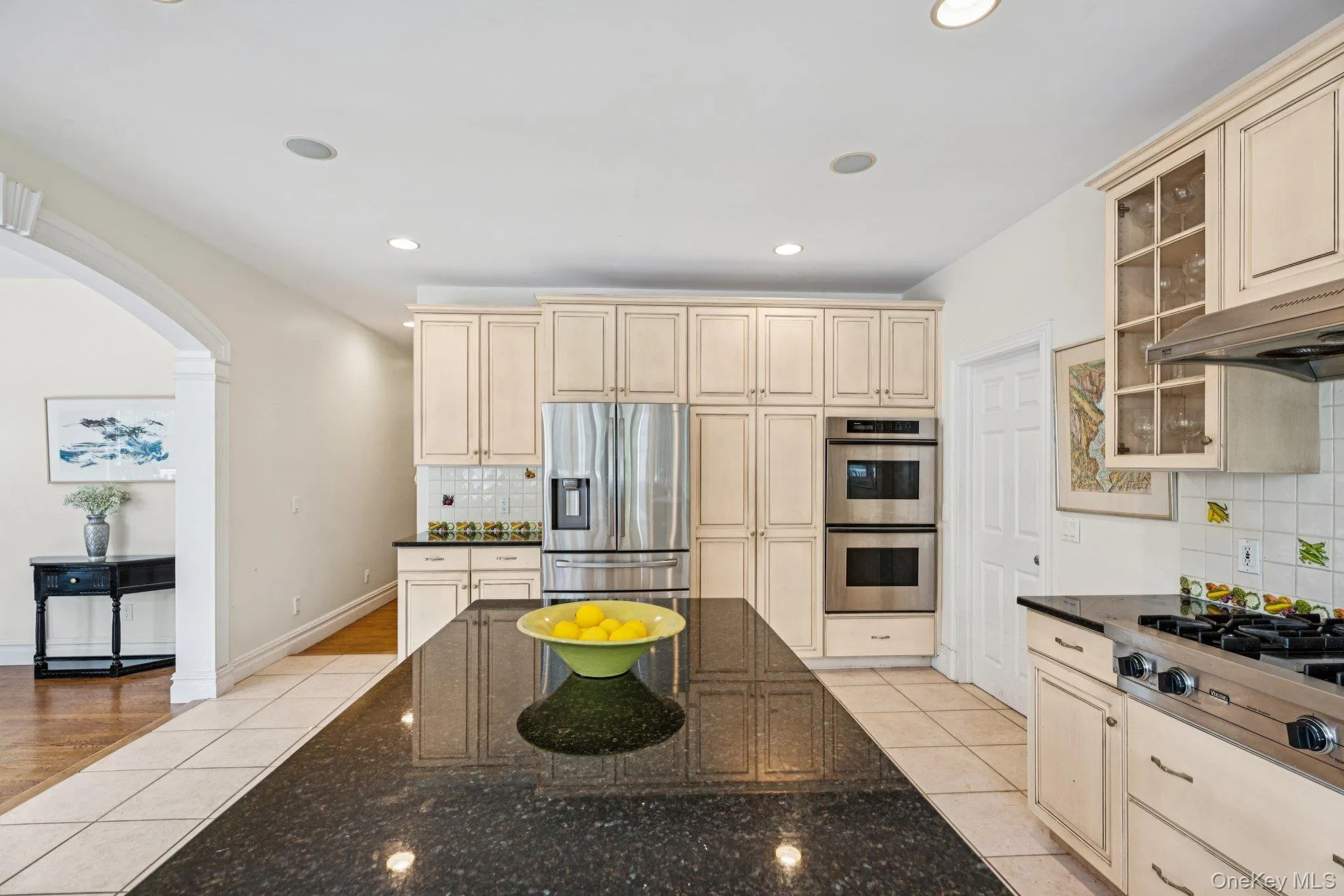 Kitchen featuring granite countertops, a spacious island, glass-inset custom cabinetry, an eat-in dining area, recessed lighting, light-patterned tile flooring. Kitchen featuring granite countertops, a spacious island, glass-inset custom cabinetry, an eat-in dining area, recessed lighting, light-patterned tile flooring.