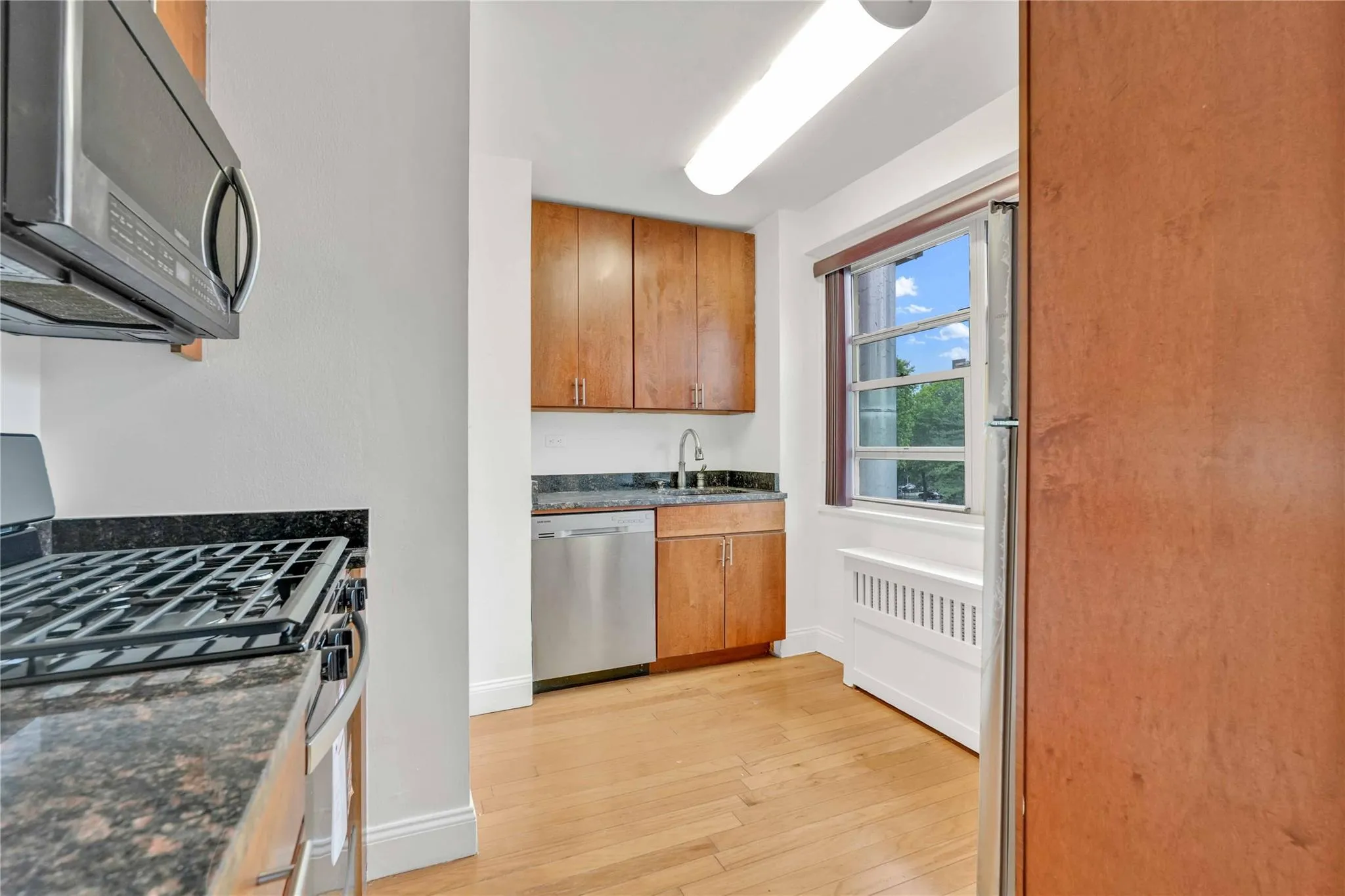 Kitchen with stainless steel appliances, light wood finished floors, radiator, and brown cabinets Kitchen with stainless steel appliances, light wood finished floors, radiator, and brown cabinets