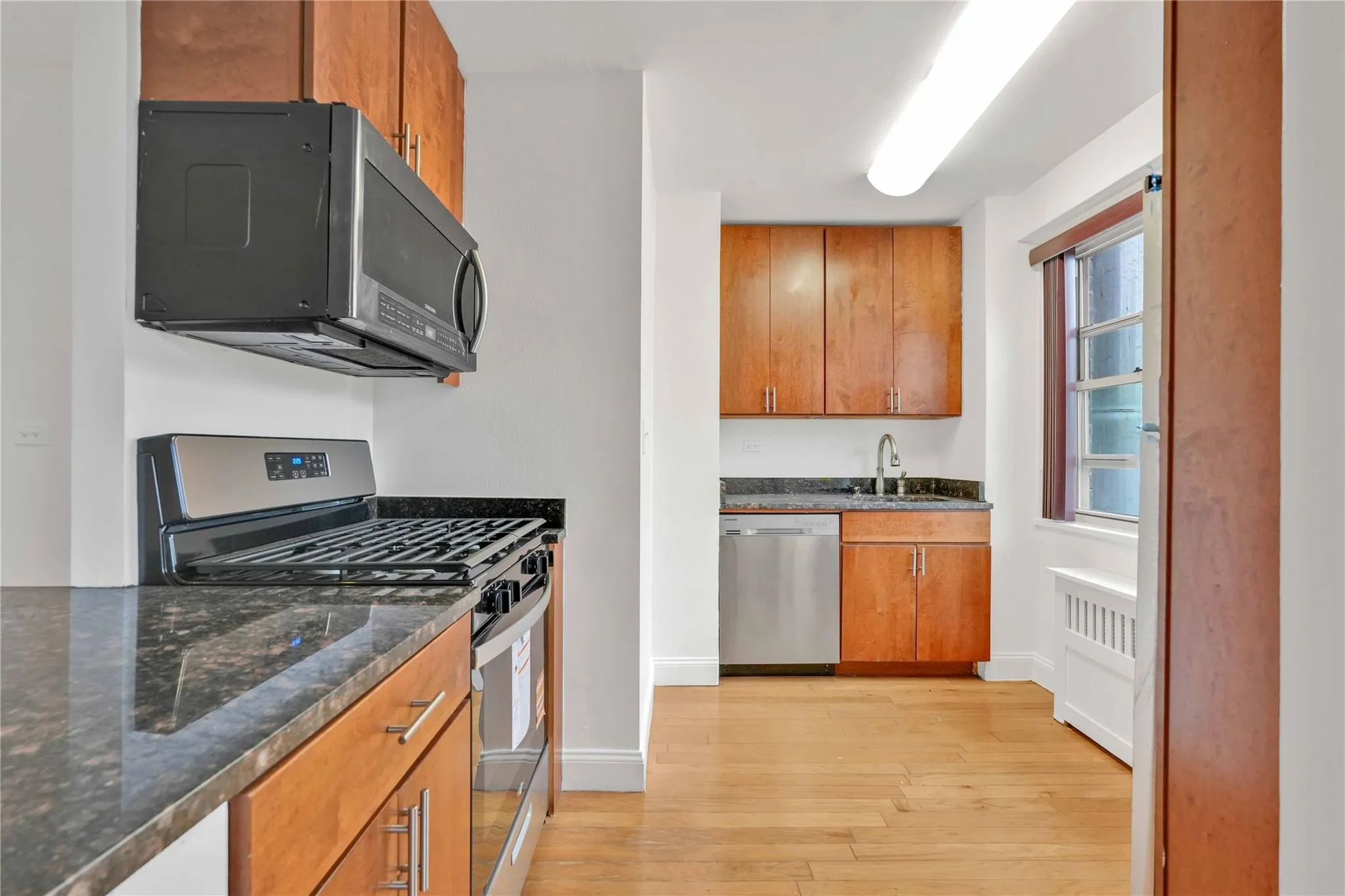 Kitchen with appliances with stainless steel finishes, light wood-type flooring, brown cabinets, dark stone counters, and radiator heating unit Kitchen with appliances with stainless steel finishes, light wood-type flooring, brown cabinets, dark stone counters, and radiator heating unit