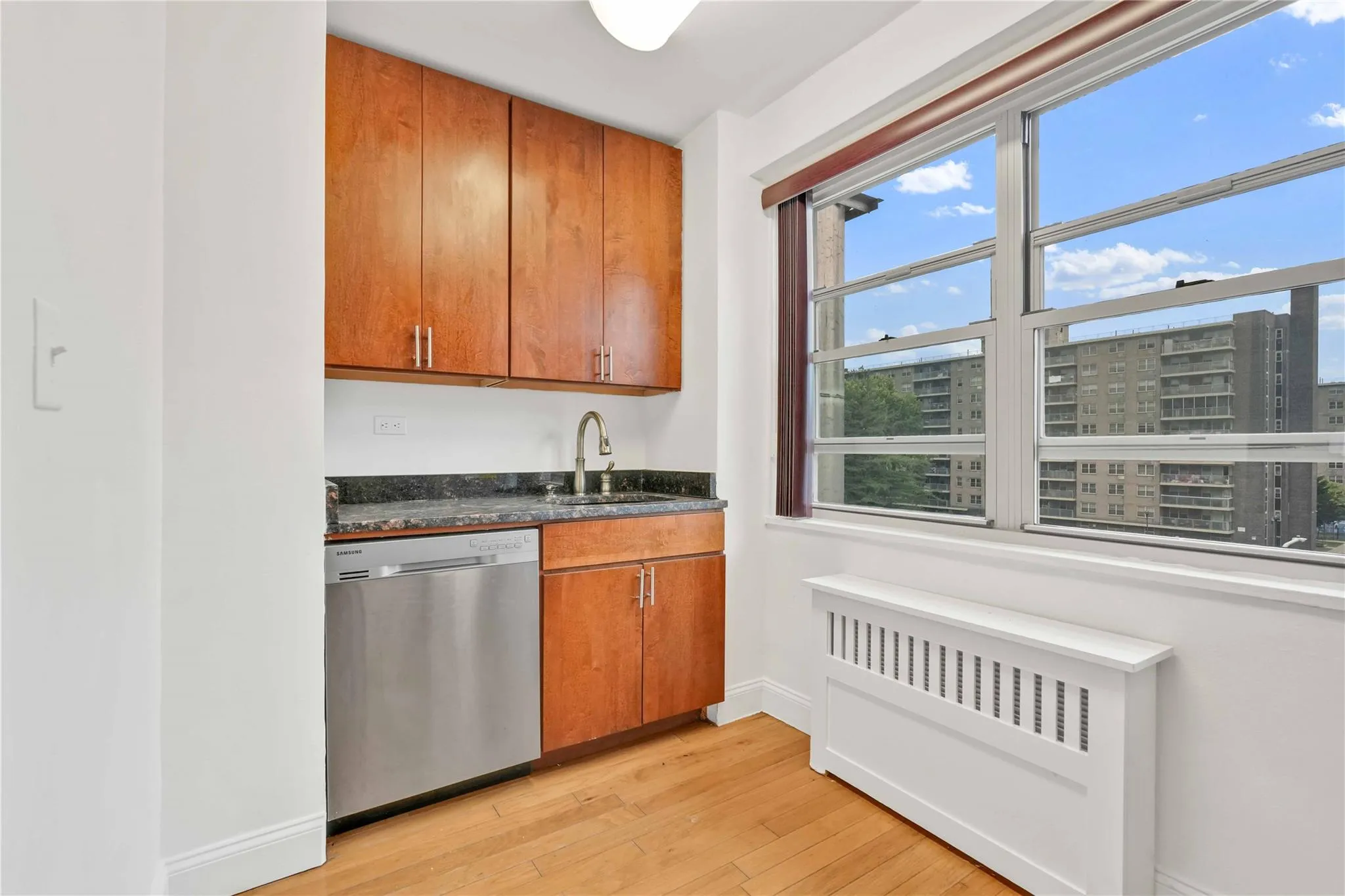 Kitchen featuring radiator heating unit, stainless steel dishwasher, light wood finished floors, brown cabinetry, and dark stone counters Kitchen featuring radiator heating unit, stainless steel dishwasher, light wood finished floors, brown cabinetry, and dark stone counters