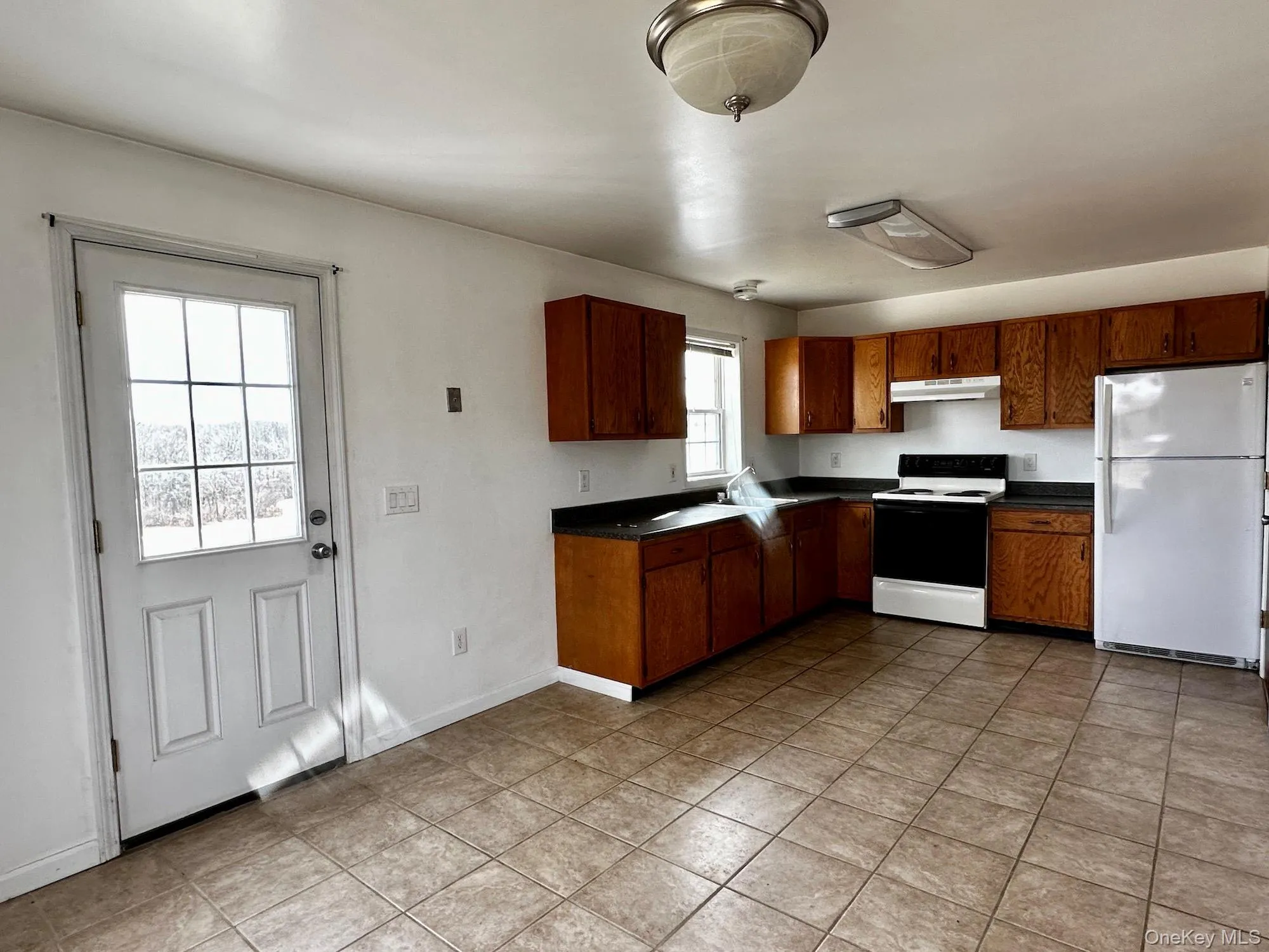 Kitchen featuring dark countertops, white appliances, under cabinet range hood, and brown cabinetry Kitchen featuring dark countertops, white appliances, under cabinet range hood, and brown cabinetry