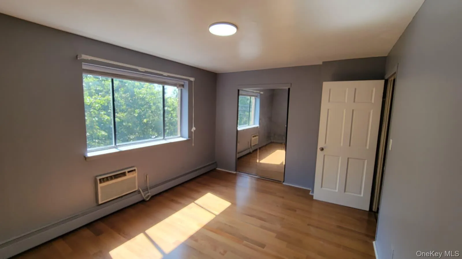 Unfurnished bedroom featuring baseboard heating, light wood finished floors, a closet, and an AC wall unit Unfurnished bedroom featuring baseboard heating, light wood finished floors, a closet, and an AC wall unit
