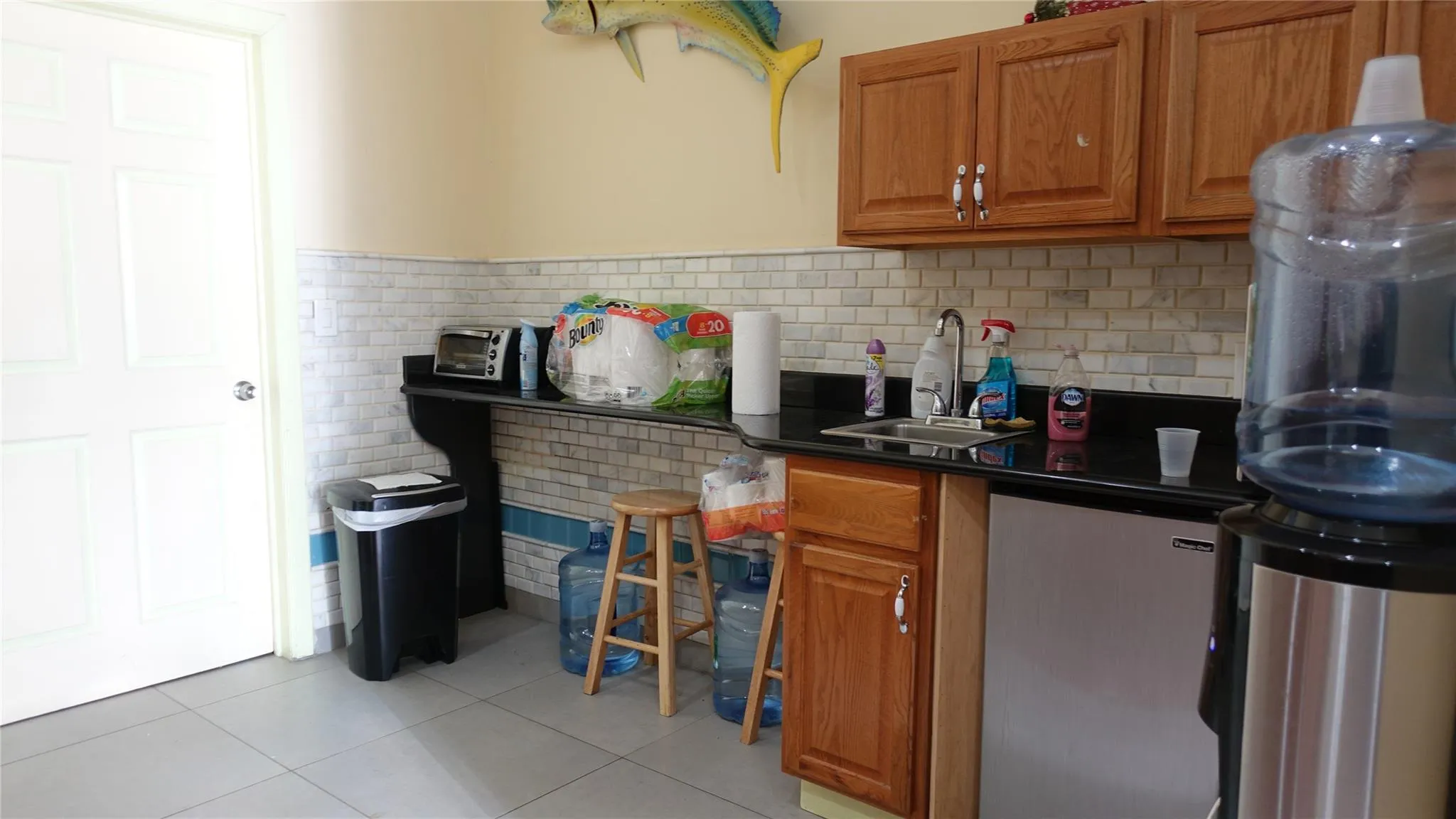 Kitchen featuring light tile patterned floors, dark countertops, brown cabinetry, and dishwashing machine Kitchen featuring light tile patterned floors, dark countertops, brown cabinetry, and dishwashing machine