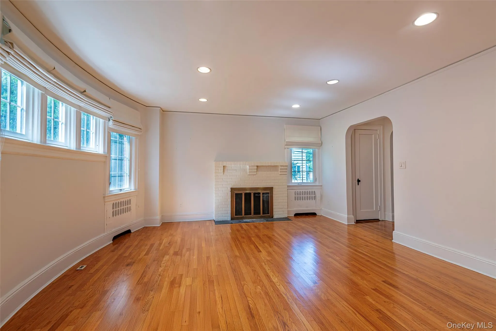 Unfurnished living room with arched walkways, light wood-type flooring, a fireplace, recessed lighting, and radiator heating unit Unfurnished living room with arched walkways, light wood-type flooring, a fireplace, recessed lighting, and radiator heating unit