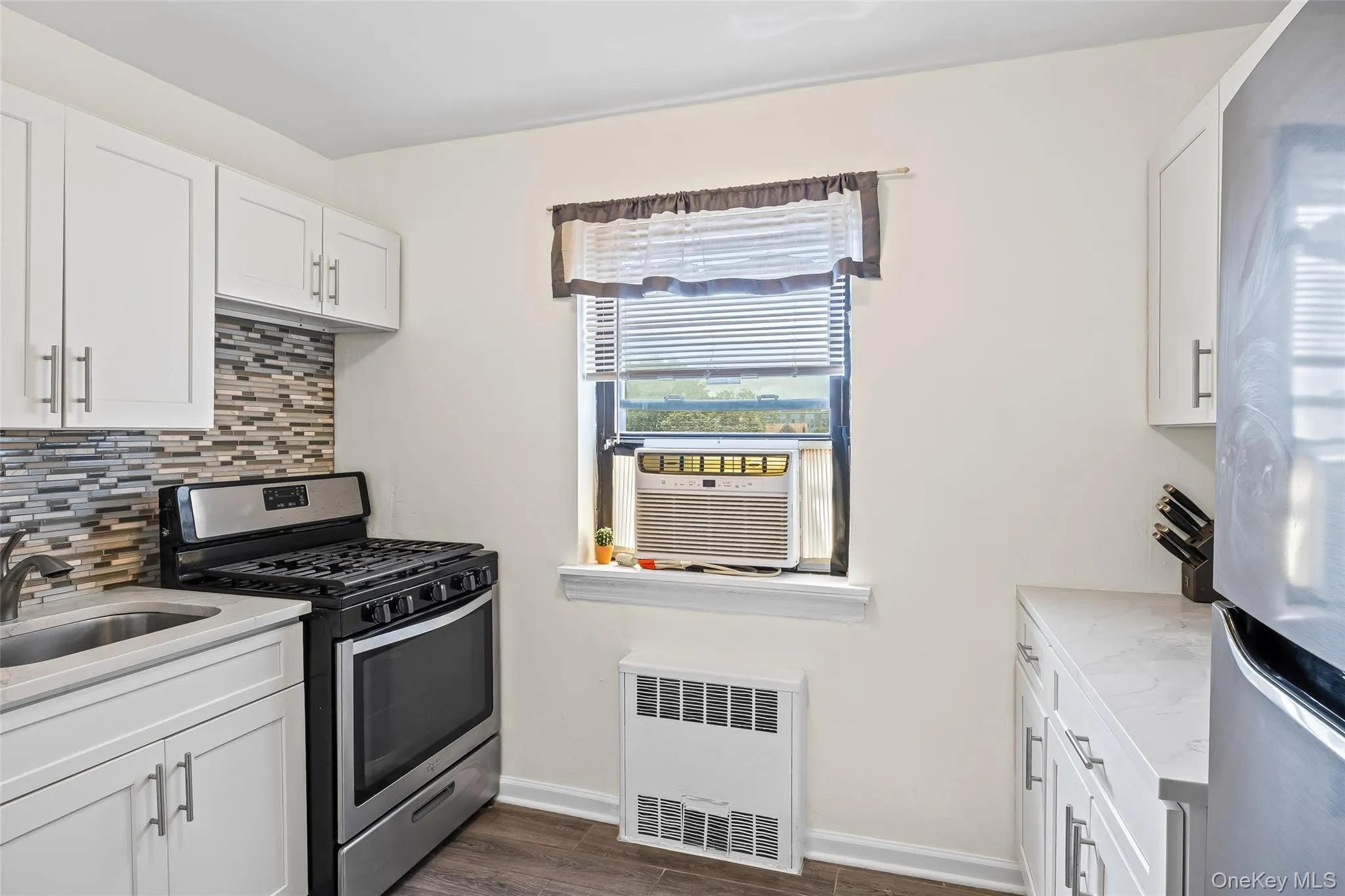 Kitchen featuring appliances with stainless steel finishes, tasteful backsplash, radiator, dark wood-type flooring, and white cabinets Kitchen featuring appliances with stainless steel finishes, tasteful backsplash, radiator, dark wood-type flooring, and white cabinets