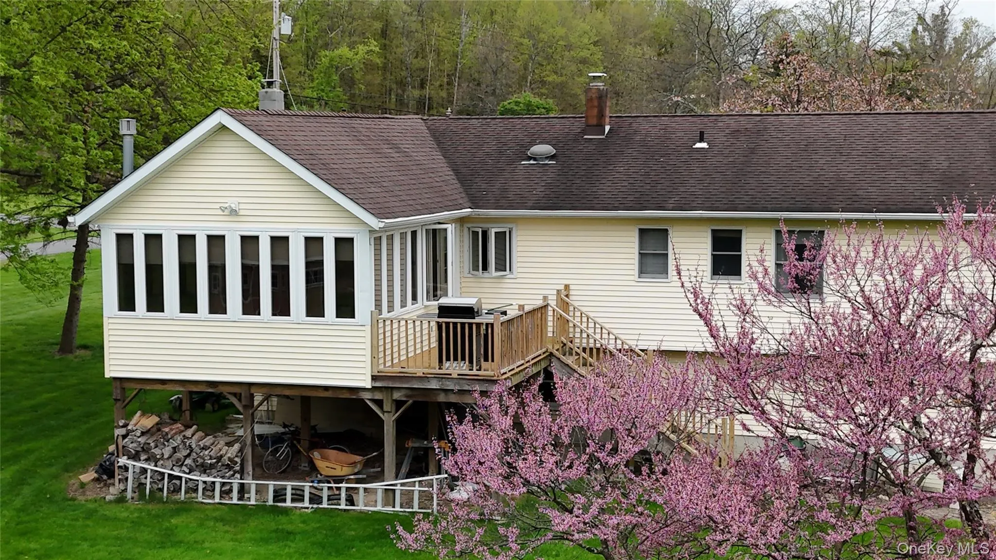 Rear exterior featuring a sunroom over storage space, an elevated wood deck with stairs, and flowering trees Rear exterior featuring a sunroom over storage space, an elevated wood deck with stairs, and flowering trees