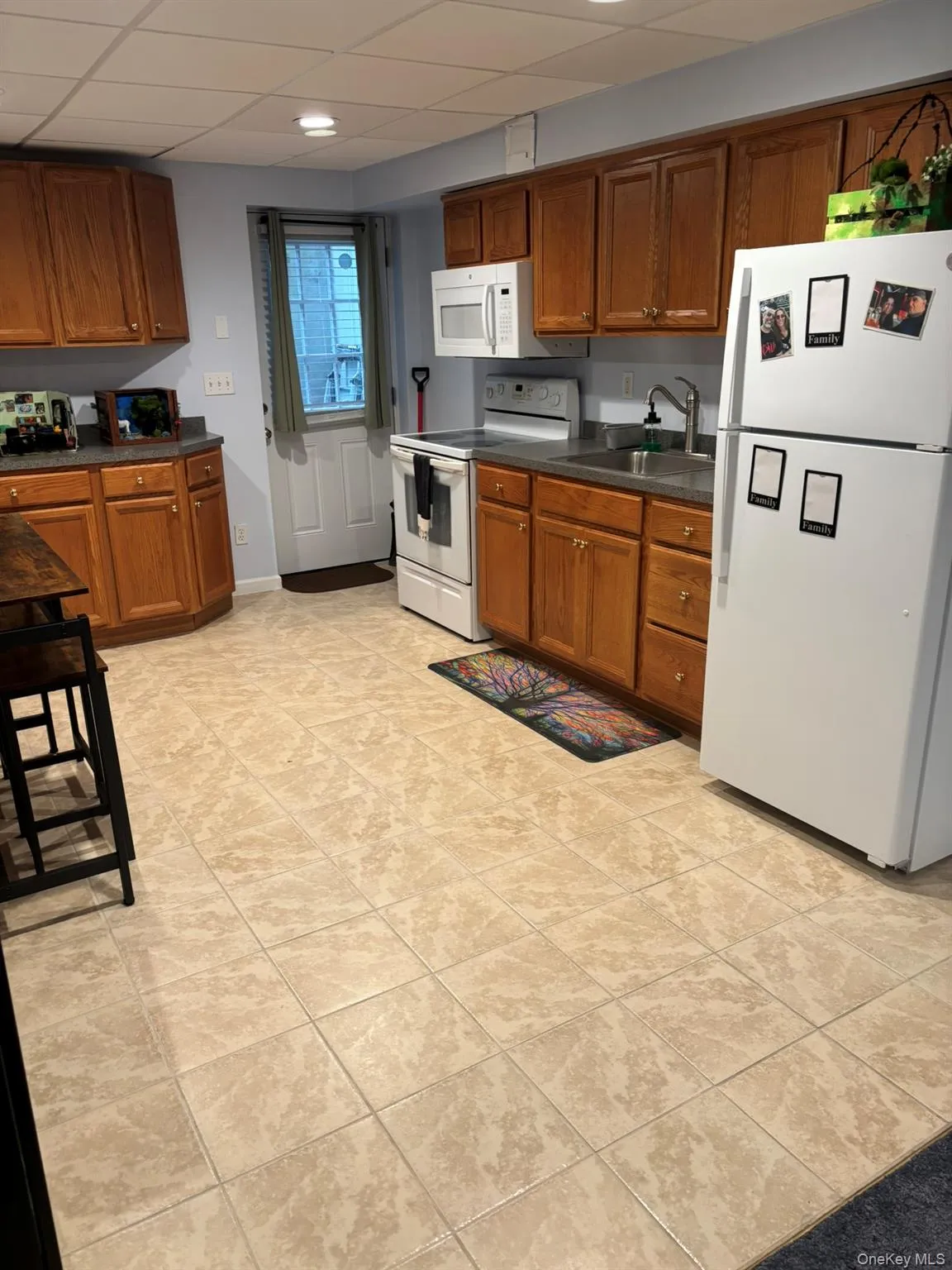 Kitchen with white appliances, a paneled ceiling, dark countertops, brown cabinetry, and light tile patterned floors Kitchen with white appliances, a paneled ceiling, dark countertops, brown cabinetry, and light tile patterned floors