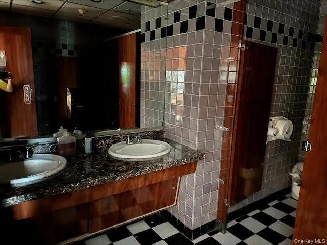 Full bath featuring tile walls, double vanity, a drop ceiling, and dark floors Full bath featuring tile walls, double vanity, a drop ceiling, and dark floors
