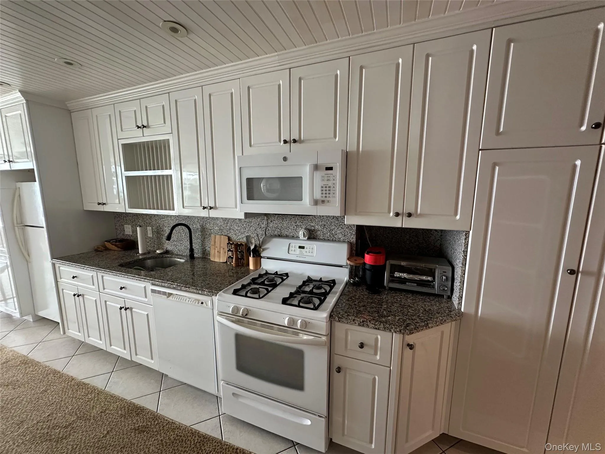 Kitchen with white appliances, light tile patterned floors, tasteful backsplash, dark stone counters, and white cabinets Kitchen with white appliances, light tile patterned floors, tasteful backsplash, dark stone counters, and white cabinets