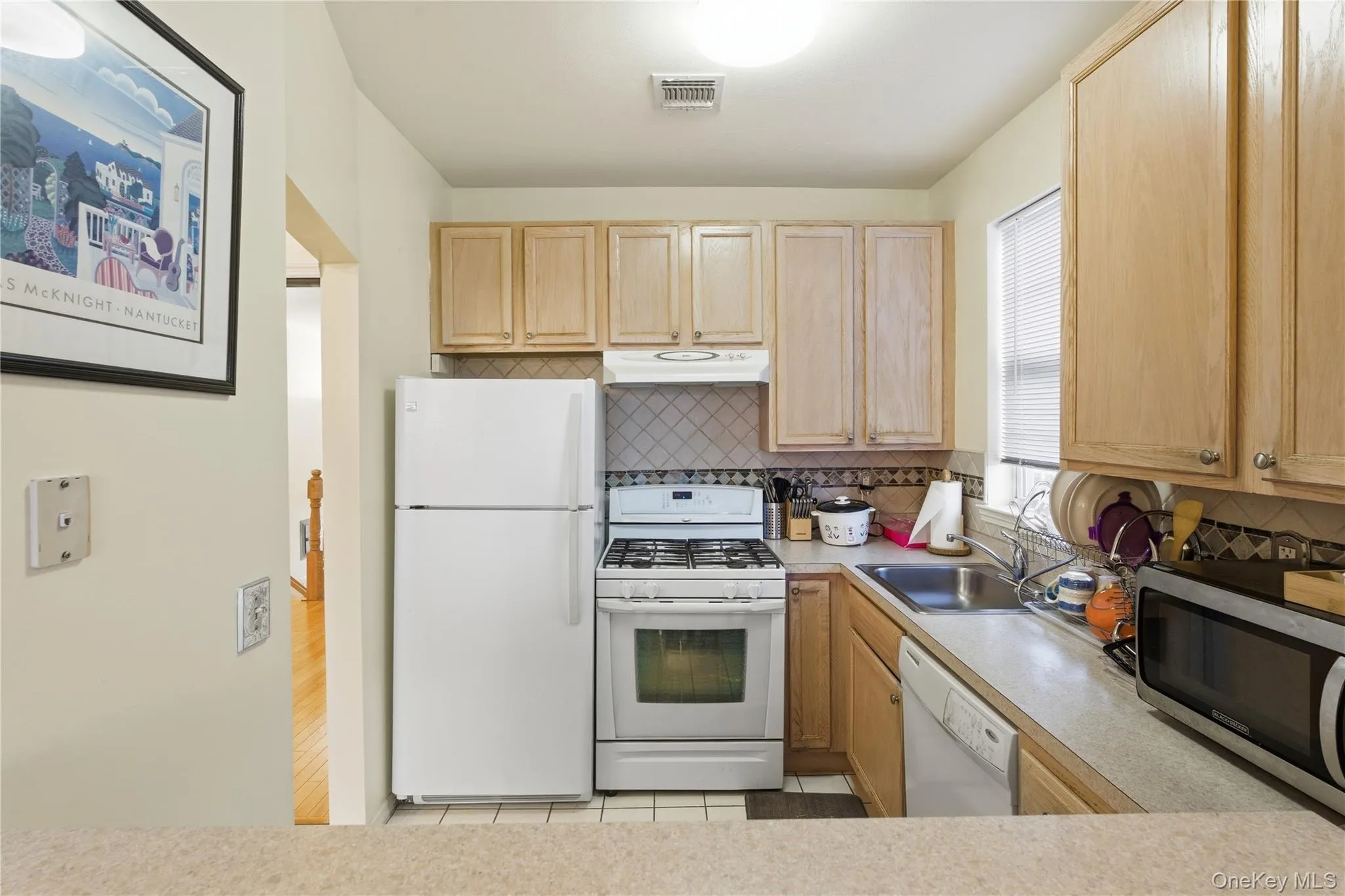 Kitchen with light brown cabinetry, white appliances, backsplash, light countertops, and under cabinet range hood Kitchen with light brown cabinetry, white appliances, backsplash, light countertops, and under cabinet range hood