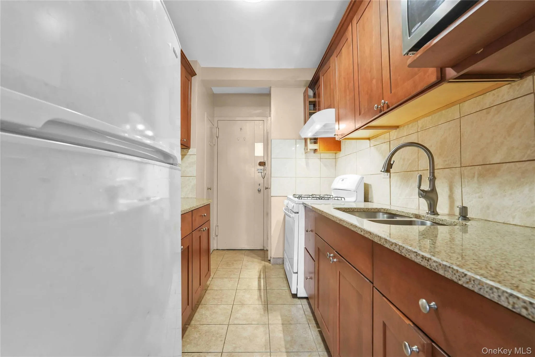 Kitchen with white appliances, light stone counters, brown cabinetry, backsplash, and ventilation hood Kitchen with white appliances, light stone counters, brown cabinetry, backsplash, and ventilation hood