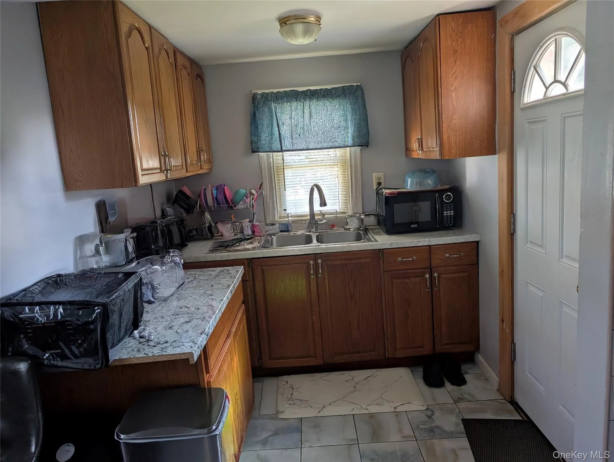 Kitchen featuring light countertops, light marble finish floors, black microwave, and brown cabinets Kitchen featuring light countertops, light marble finish floors, black microwave, and brown cabinets
