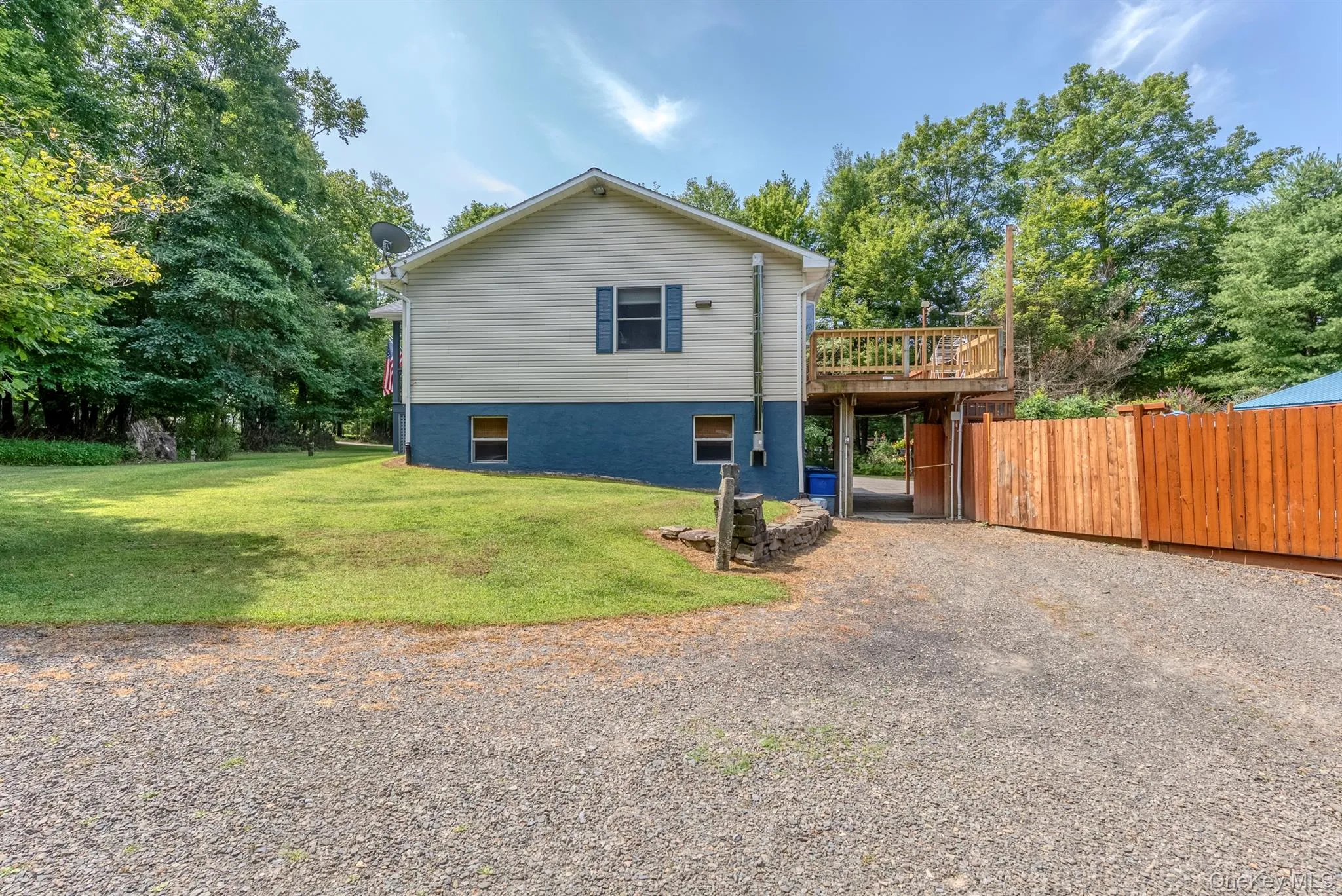 Rear view of property with a wooden deck, a lawn, gravel driveway, and view of scattered trees Rear view of property with a wooden deck, a lawn, gravel driveway, and view of scattered trees