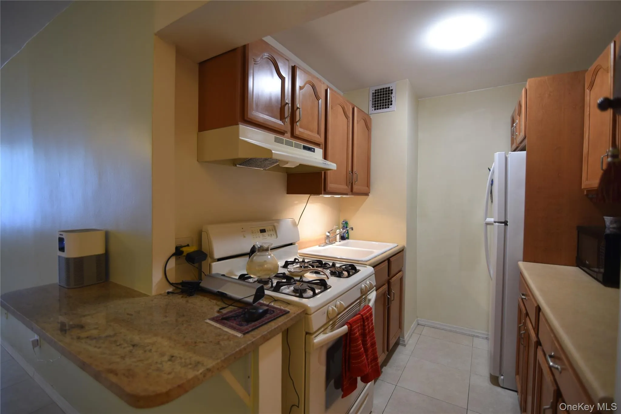 Kitchen featuring white appliances, under cabinet range hood, brown cabinetry, and light tile patterned floors Kitchen featuring white appliances, under cabinet range hood, brown cabinetry, and light tile patterned floors