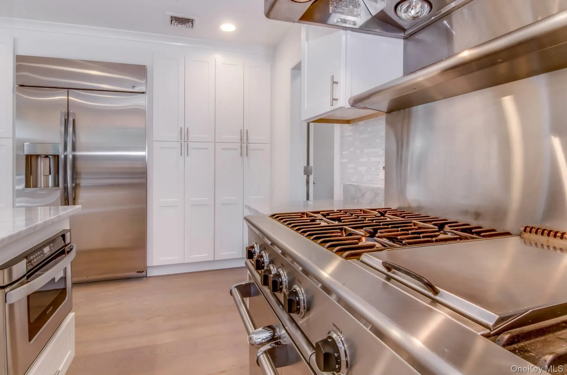 Kitchen with appliances with stainless steel finishes, white cabinets, backsplash, light wood-style flooring, and light stone countertops Kitchen with appliances with stainless steel finishes, white cabinets, backsplash, light wood-style flooring, and light stone countertops