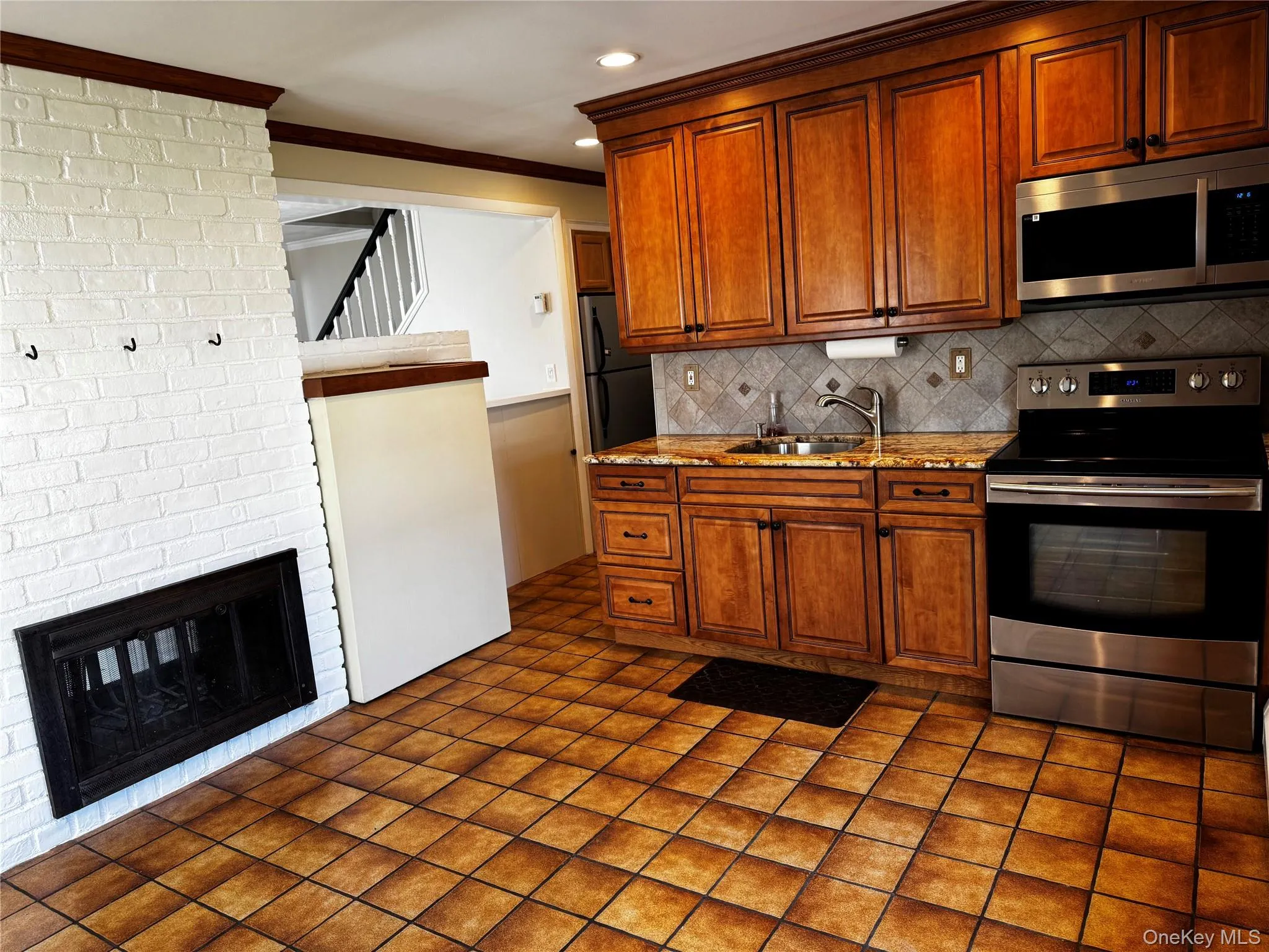 Kitchen with stainless steel appliances, brown cabinetry, light stone countertops, a brick fireplace, and tasteful backsplash Kitchen with stainless steel appliances, brown cabinetry, light stone countertops, a brick fireplace, and tasteful backsplash