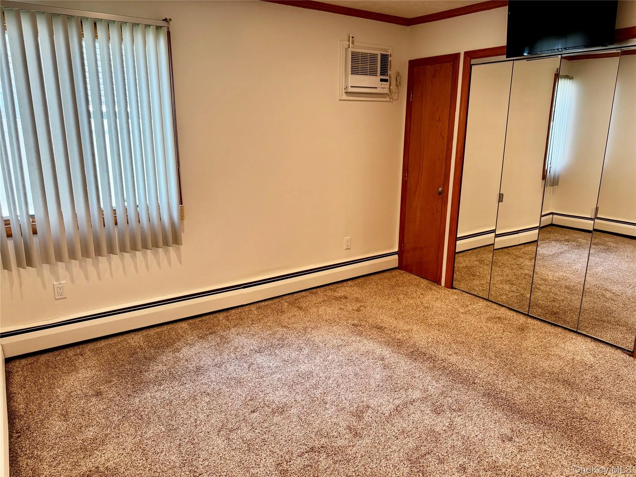 Unfurnished bedroom with baseboard heating, a closet, carpet, ornamental molding, and a wall unit AC Unfurnished bedroom with baseboard heating, a closet, carpet, ornamental molding, and a wall unit AC