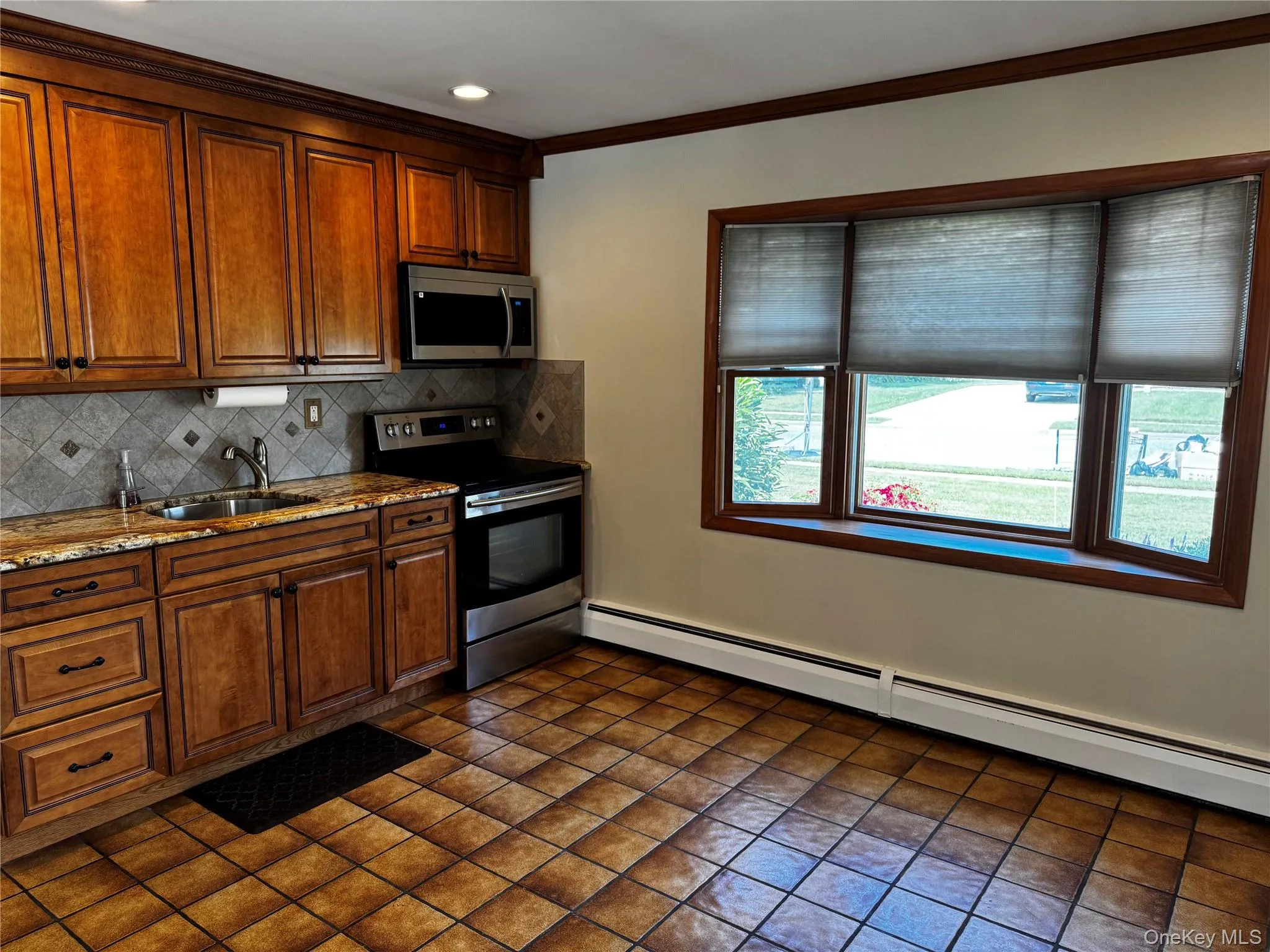Kitchen featuring stainless steel appliances, light stone counters, baseboard heating, tasteful backsplash, and brown cabinetry Kitchen featuring stainless steel appliances, light stone counters, baseboard heating, tasteful backsplash, and brown cabinetry