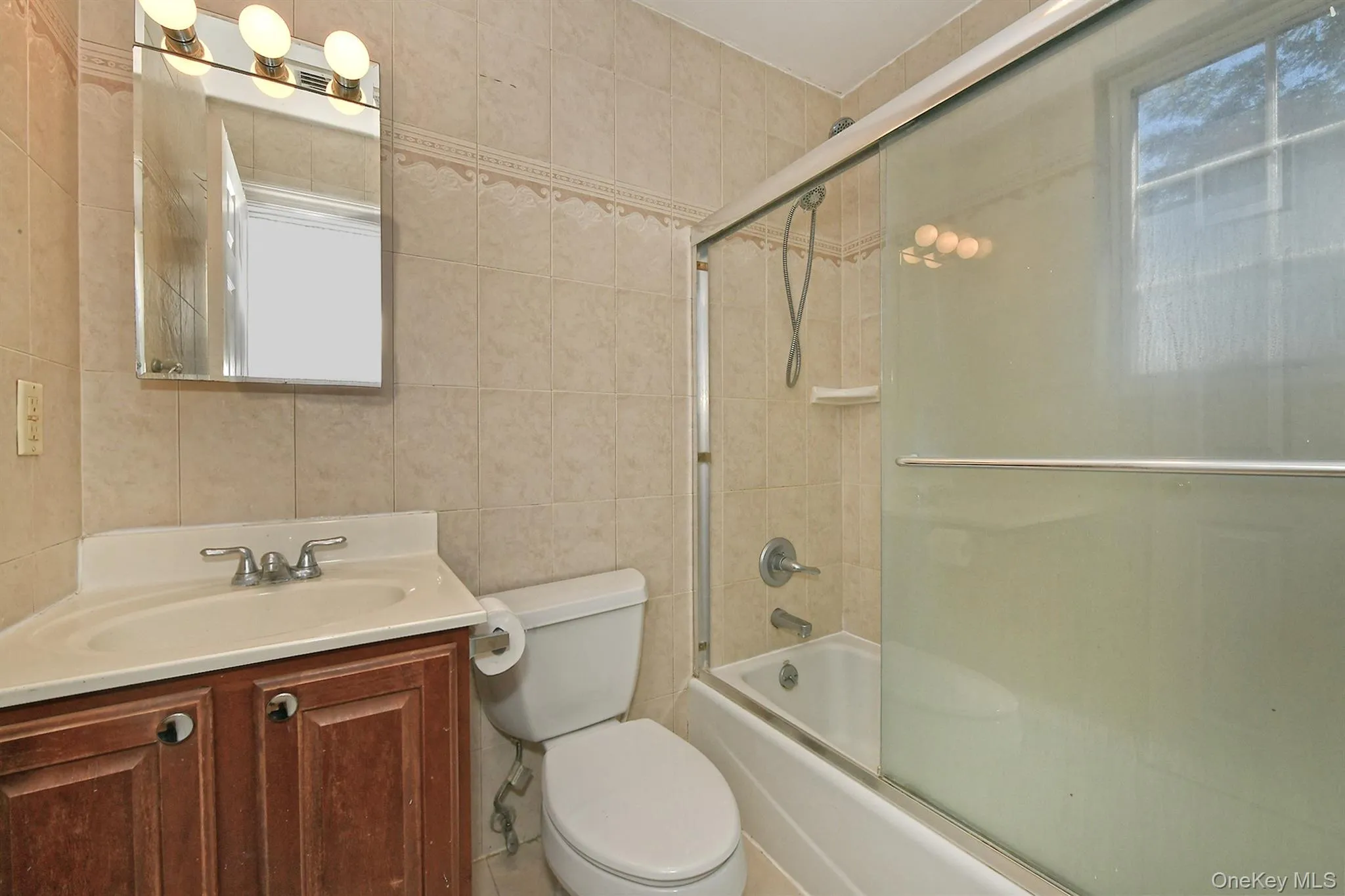Full bathroom with tile walls, shower / bath combination with glass door, healthy amount of natural light, and vanity Full bathroom with tile walls, shower / bath combination with glass door, healthy amount of natural light, and vanity