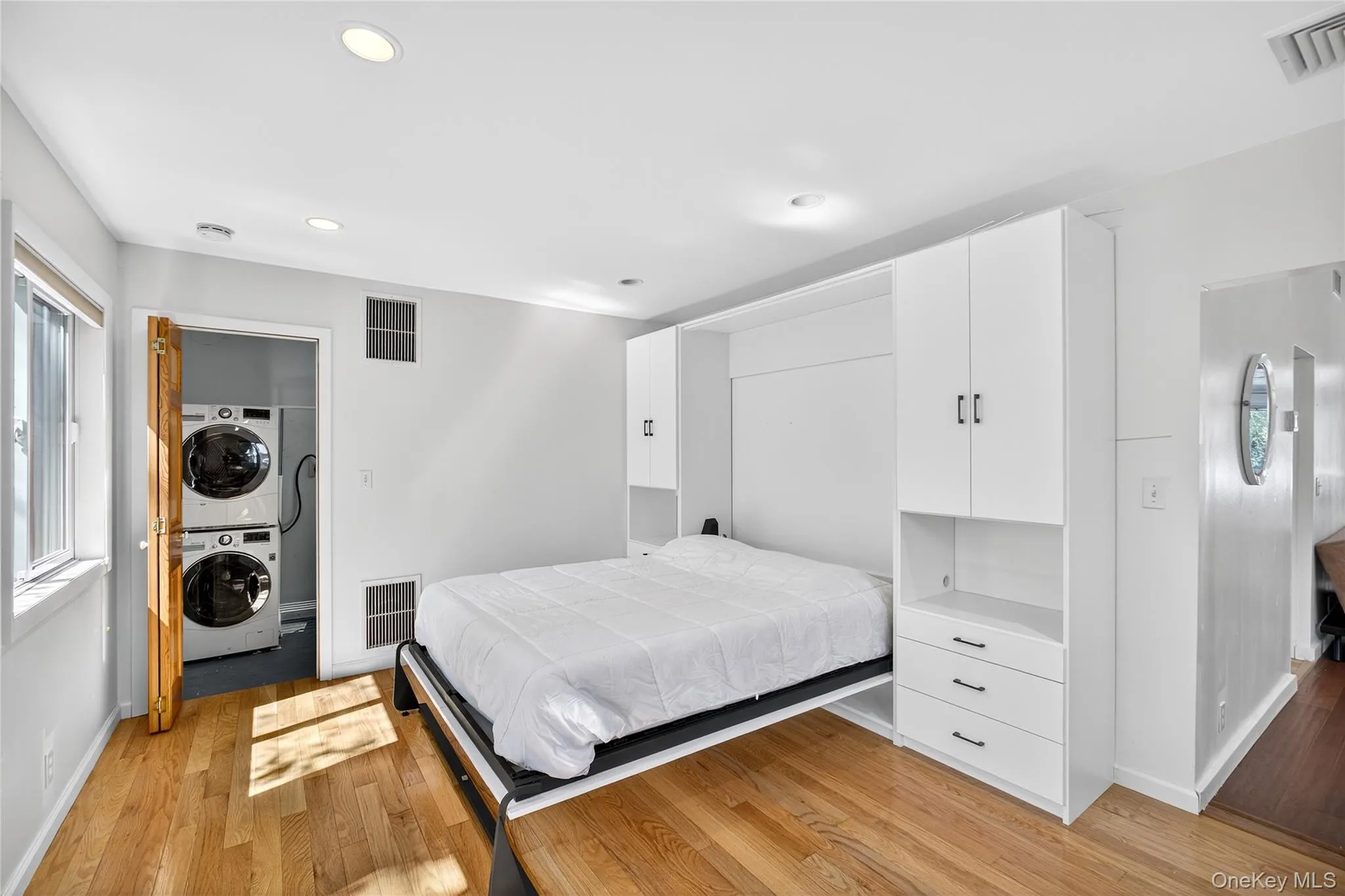 Primary Bedroom with Murphy bed or can be closed and used as a wall, stackable washer and clothes dryer in separate laundry room as seen light wood-type flooring, and recessed lighting Primary Bedroom with Murphy bed or can be closed and used as a wall, stackable washer and clothes dryer in separate laundry room as seen light wood-type flooring, and recessed lighting