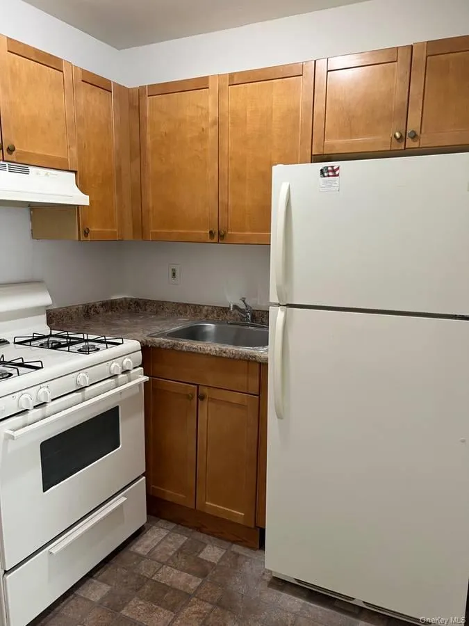 Kitchen featuring white appliances, brown cabinetry, under cabinet range hood, stone finish flooring, and dark countertops Kitchen featuring white appliances, brown cabinetry, under cabinet range hood, stone finish flooring, and dark countertops