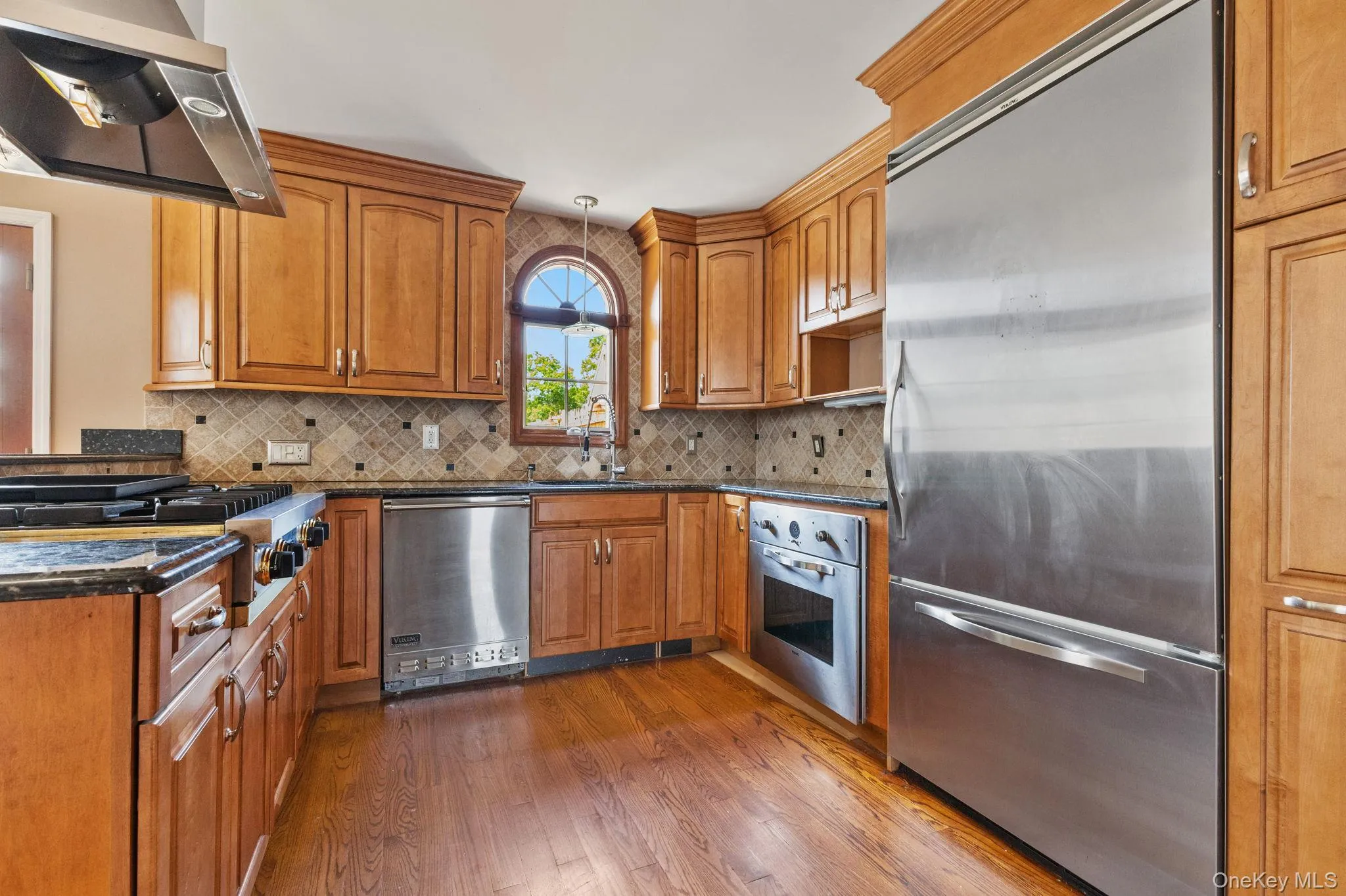 Kitchen with appliances with stainless steel finishes, brown cabinetry, dark stone countertops, range hood, and dark wood-type flooring Kitchen with appliances with stainless steel finishes, brown cabinetry, dark stone countertops, range hood, and dark wood-type flooring