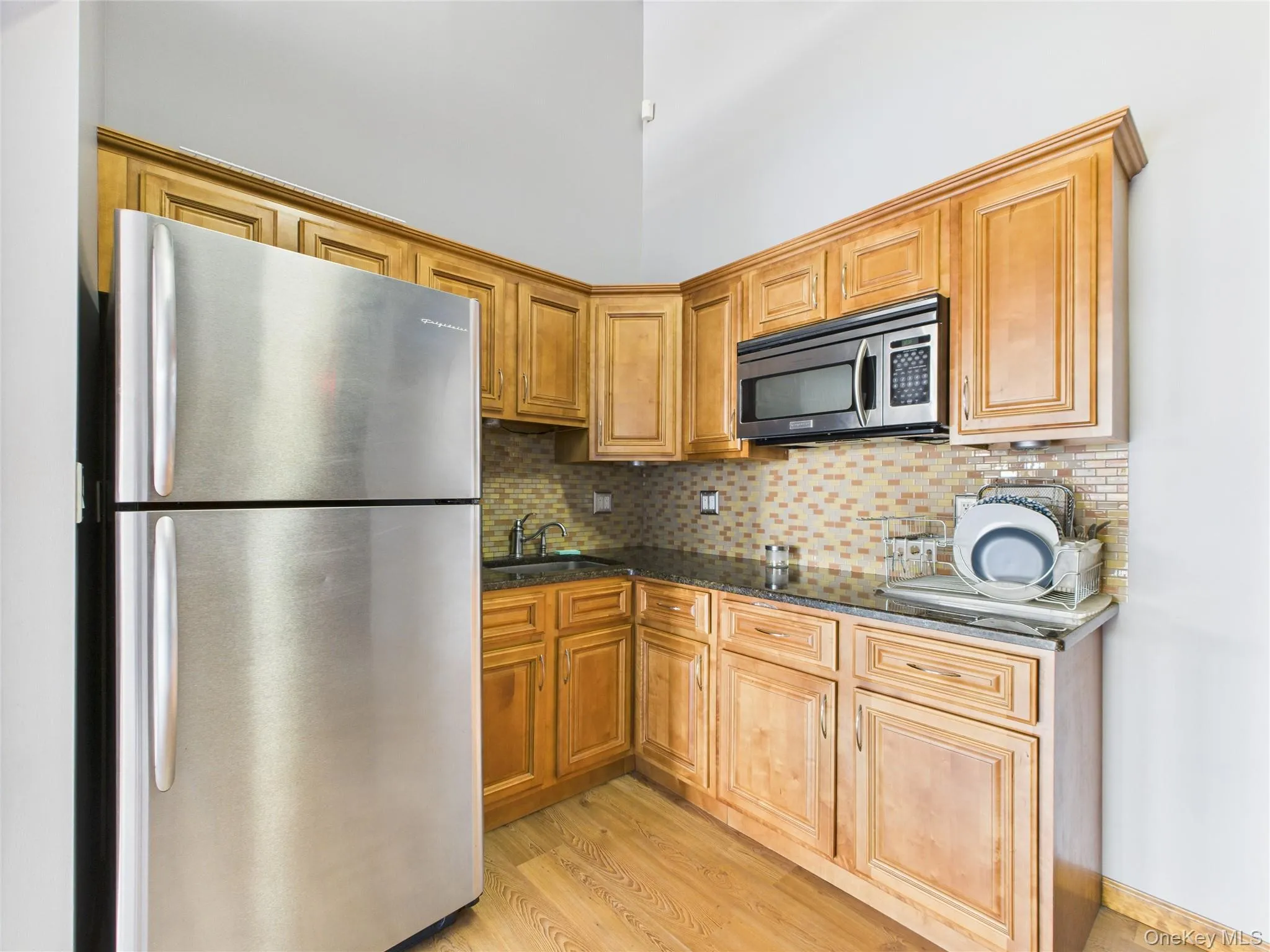 Kitchen with stainless steel appliances, decorative backsplash, light wood-type flooring, dark stone counters, and brown cabinets Kitchen with stainless steel appliances, decorative backsplash, light wood-type flooring, dark stone counters, and brown cabinets