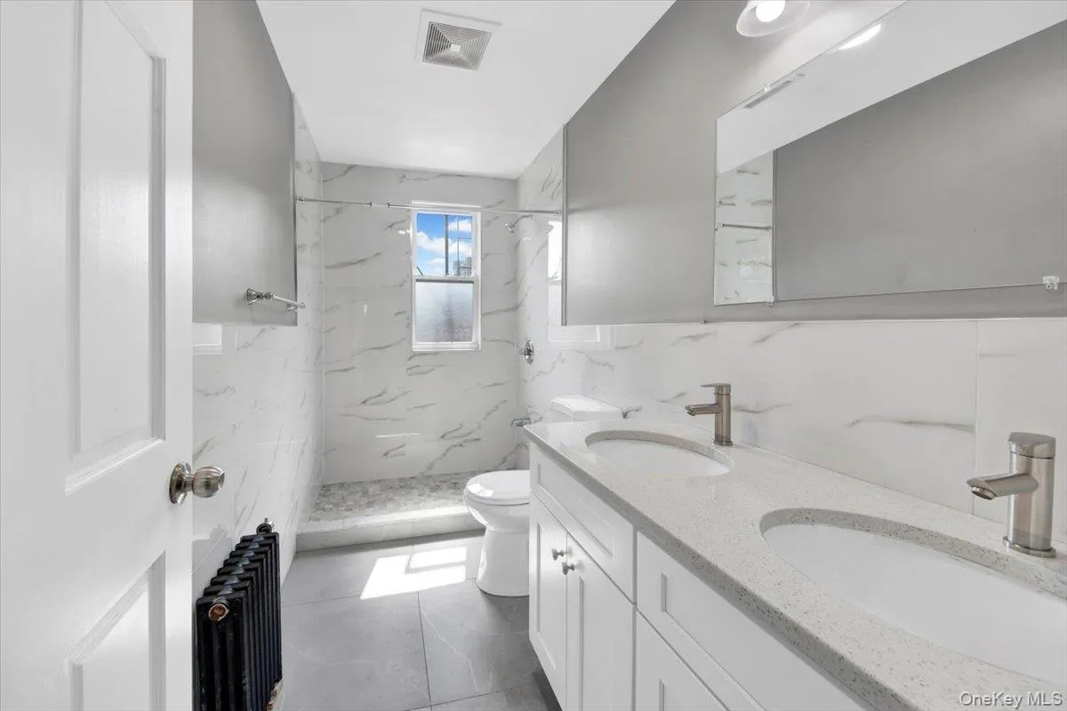 Full bath with radiator, double vanity, a marble finish shower, and tile walls Full bath with radiator, double vanity, a marble finish shower, and tile walls