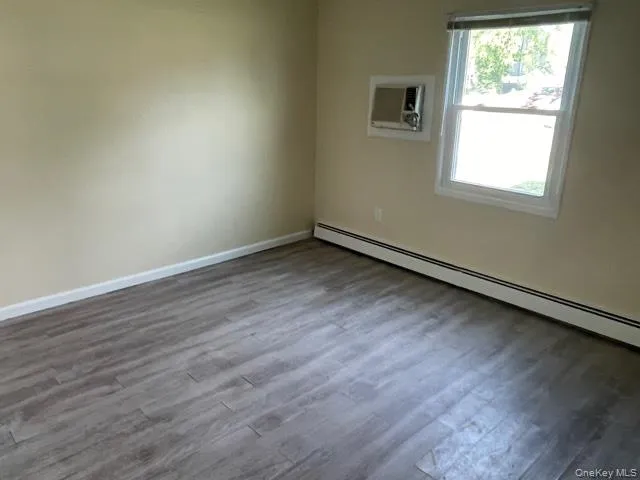 Empty room with baseboard heating, dark wood-style flooring, and an AC wall unit Empty room with baseboard heating, dark wood-style flooring, and an AC wall unit