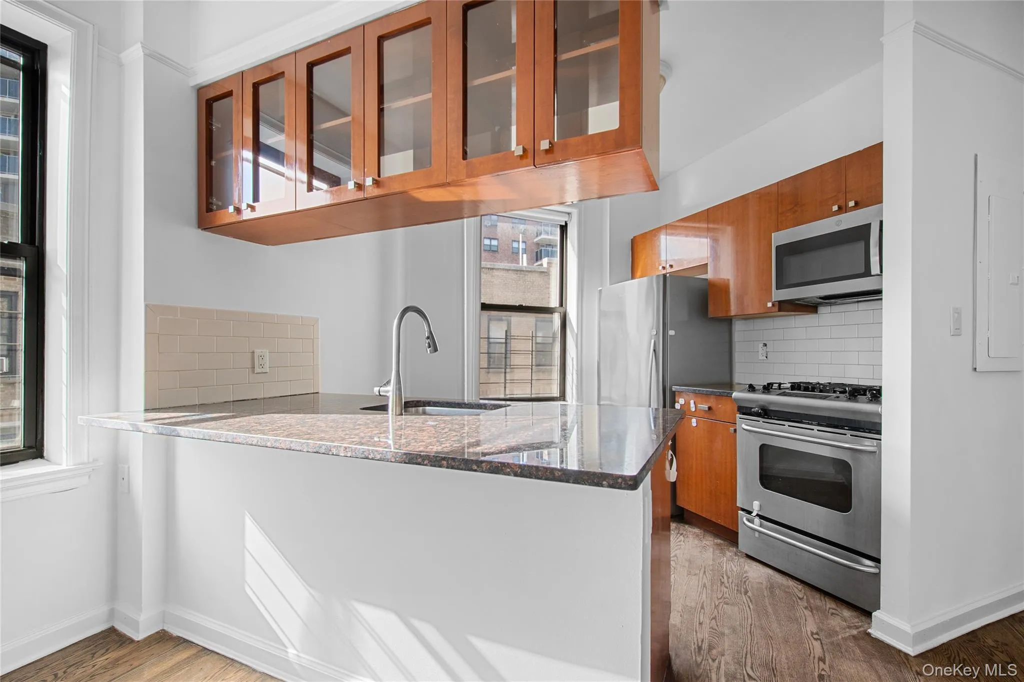 Kitchen with tasteful backsplash, brown cabinets, glass insert cabinets, dark stone countertops, and appliances with stainless steel finishes Kitchen with tasteful backsplash, brown cabinets, glass insert cabinets, dark stone countertops, and appliances with stainless steel finishes