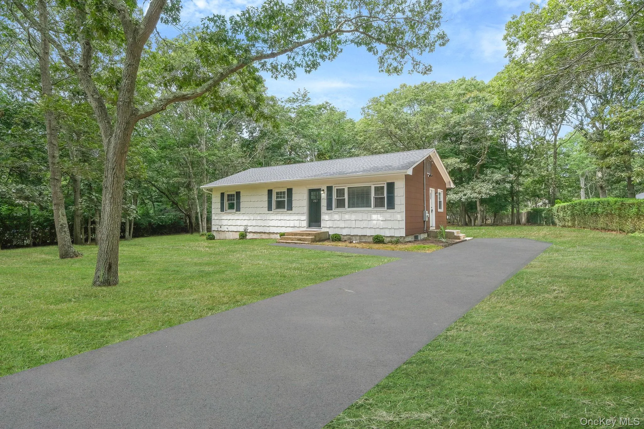 Single story home with a front yard, driveway, a shingled roof, and view of wooded area