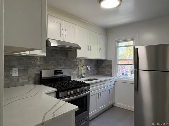 Kitchen with appliances with stainless steel finishes, backsplash, white cabinetry, under cabinet range hood, and light stone counters Kitchen with appliances with stainless steel finishes, backsplash, white cabinetry, under cabinet range hood, and light stone counters