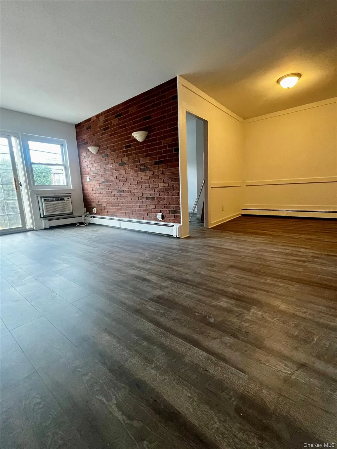 Unfurnished living room with dark wood-style floors, brick wall, a baseboard radiator, and a wall mounted AC Unfurnished living room with dark wood-style floors, brick wall, a baseboard radiator, and a wall mounted AC
