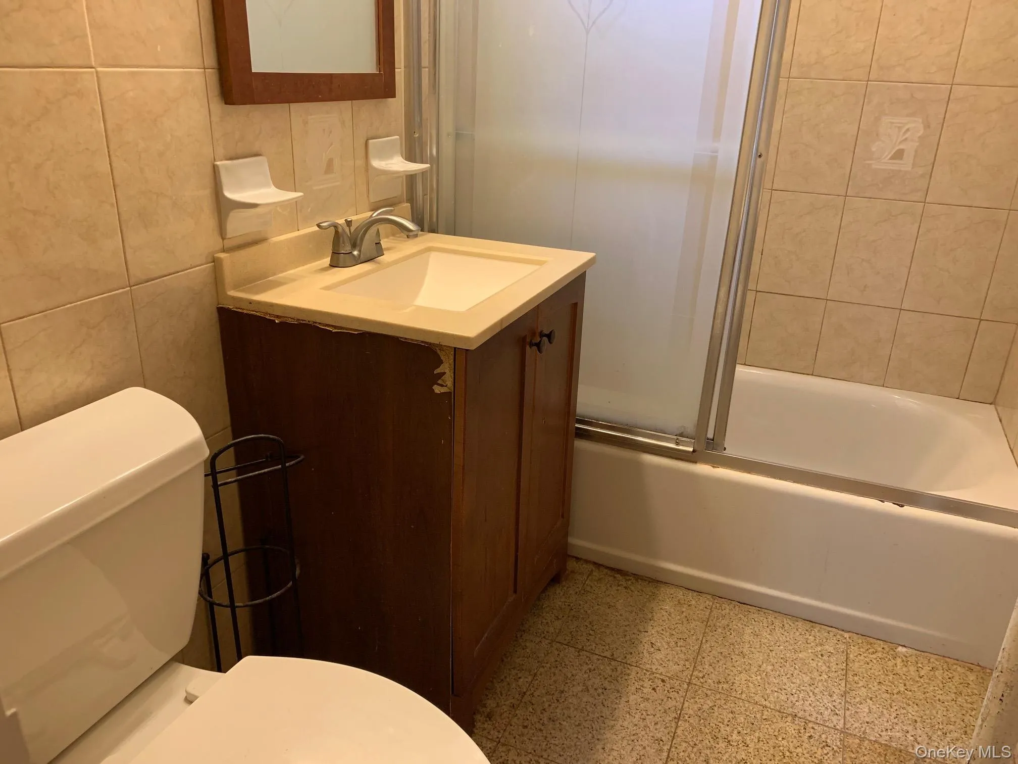 Full bathroom with tile walls, vanity, shower / bath combination with glass door, and dark speckled floor Full bathroom with tile walls, vanity, shower / bath combination with glass door, and dark speckled floor