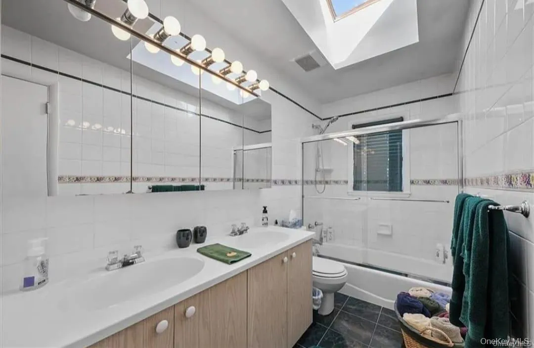 Full bath with a skylight, shower / bath combination with glass door, tile walls, double vanity, and tasteful backsplash Full bath with a skylight, shower / bath combination with glass door, tile walls, double vanity, and tasteful backsplash