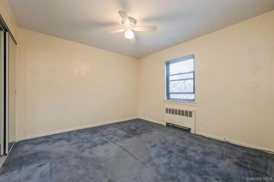 Empty room featuring radiator heating unit, dark carpet, and ceiling fan Empty room featuring radiator heating unit, dark carpet, and ceiling fan