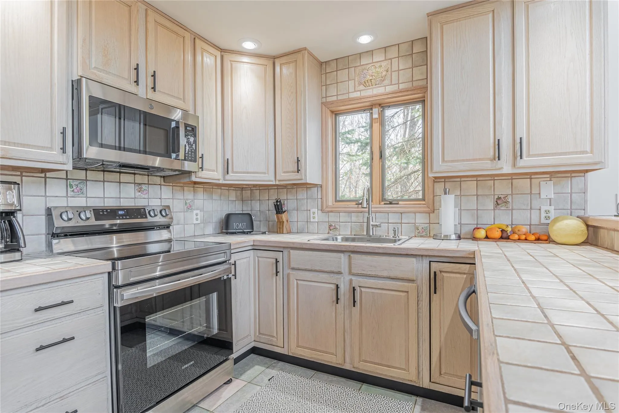 Kitchen featuring tile countertops, stainless steel appliances, light brown cabinetry, light tile patterned floors, and backsplash Kitchen featuring tile countertops, stainless steel appliances, light brown cabinetry, light tile patterned floors, and backsplash