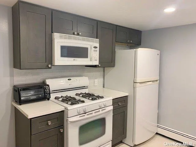 Kitchen featuring white appliances, light countertops, tasteful backsplash, a baseboard heating unit, and gray cabinetry Kitchen featuring white appliances, light countertops, tasteful backsplash, a baseboard heating unit, and gray cabinetry