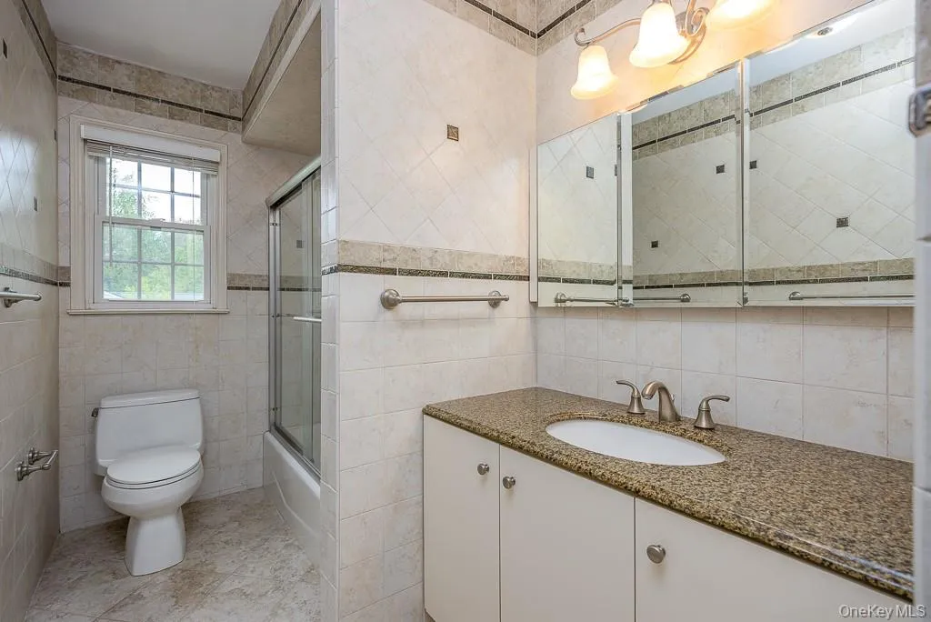 Full bath with tile walls, vanity, toilet, shower / bath combination with glass door, and tile patterned flooring Full bath with tile walls, vanity, toilet, shower / bath combination with glass door, and tile patterned flooring
